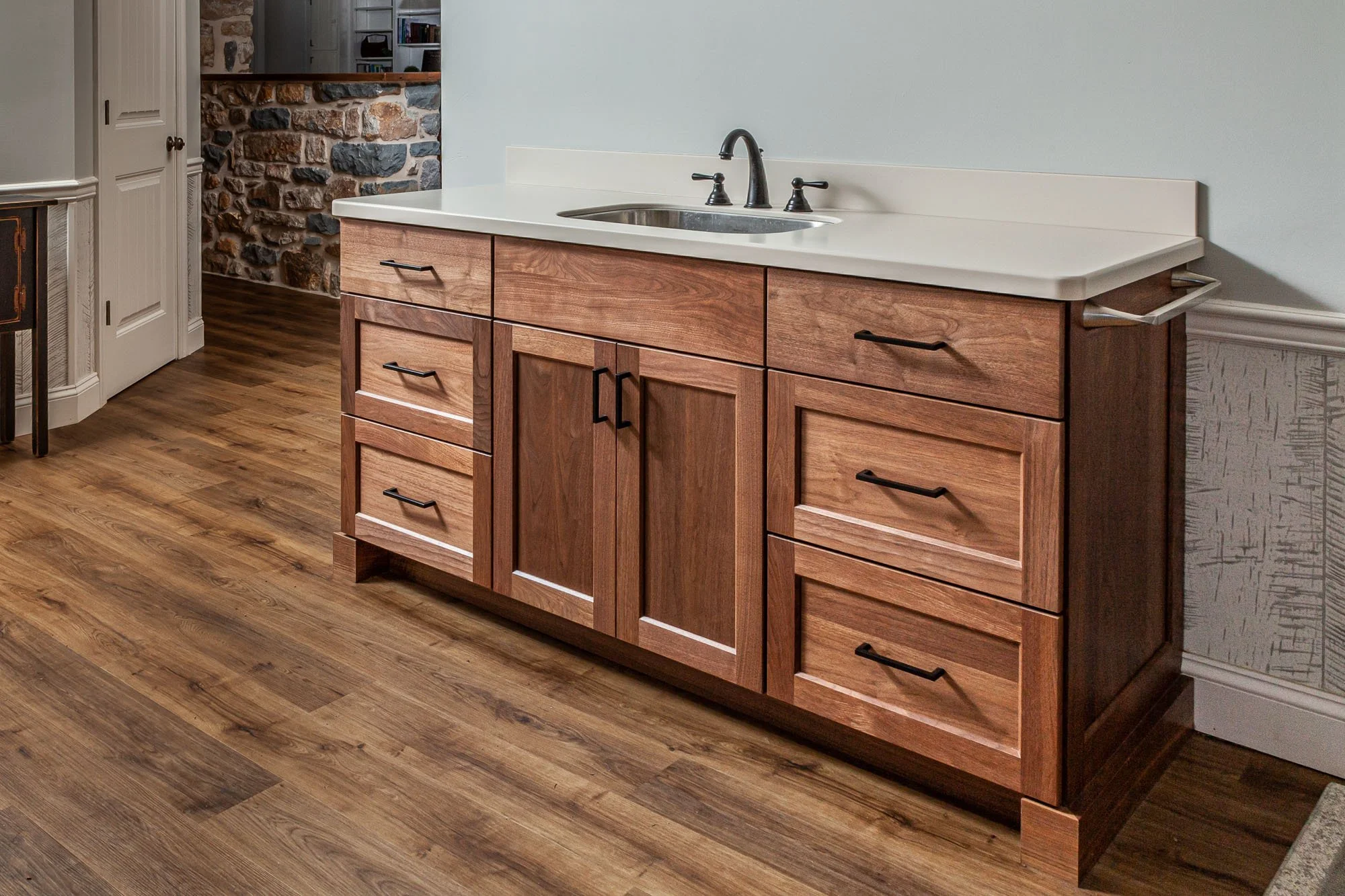 A wooden bathroom vanity with multiple drawers and cabinets, a white countertop, and a black faucet, positioned against a light-colored wall with wooden flooring.