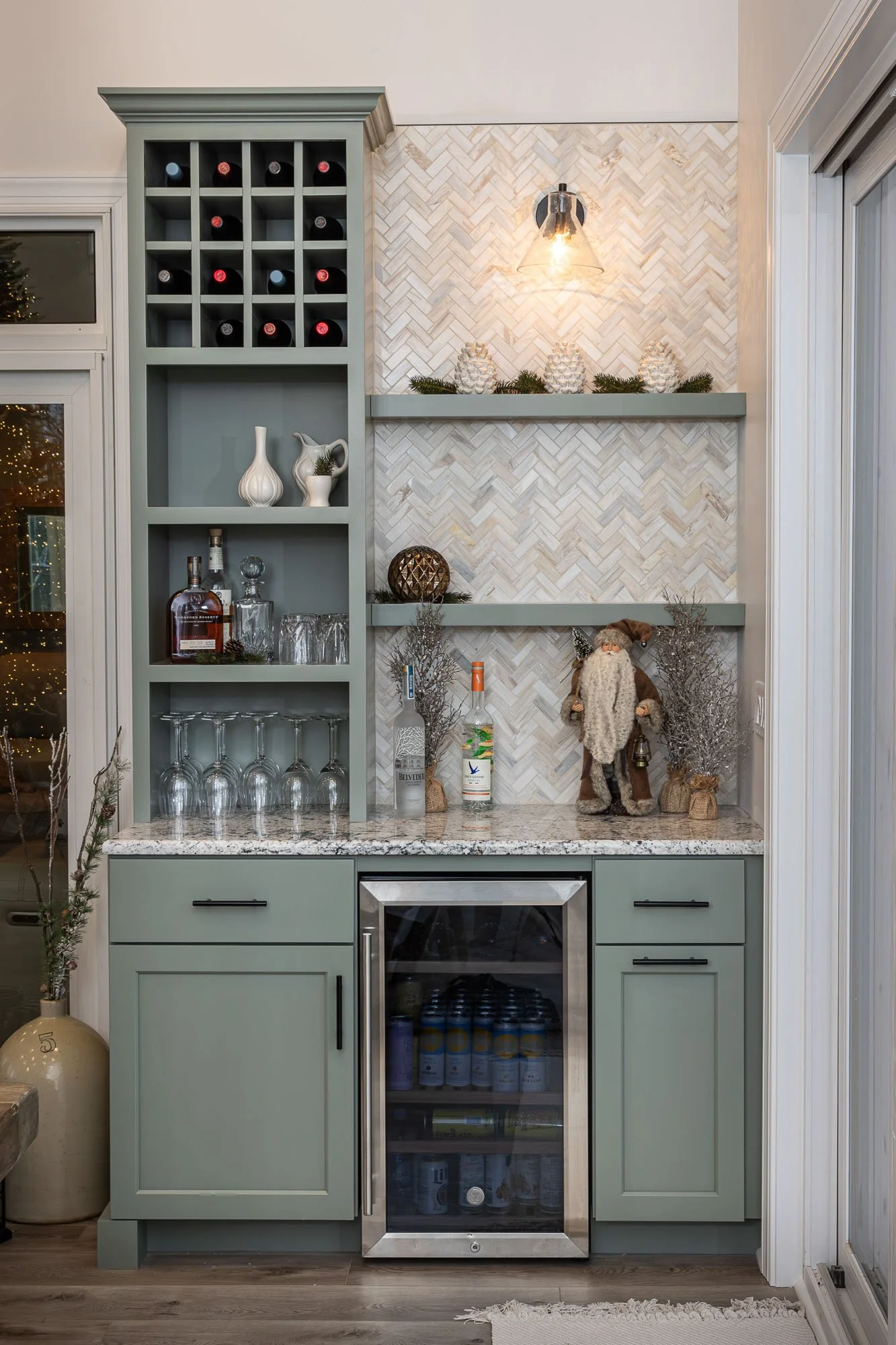 A kitchen area featuring a built-in bar or kitchenette with a teal cabinet that has a wine rack on top and shelves with decorative items. There is a small wine fridge, a countertop with bottles, glasses, and decorative items such as a Santa figure, pinecones, and greenery. The backsplash has a herringbone pattern, and there is a light fixture above. To the right, there is a sliding glass door with white trim.