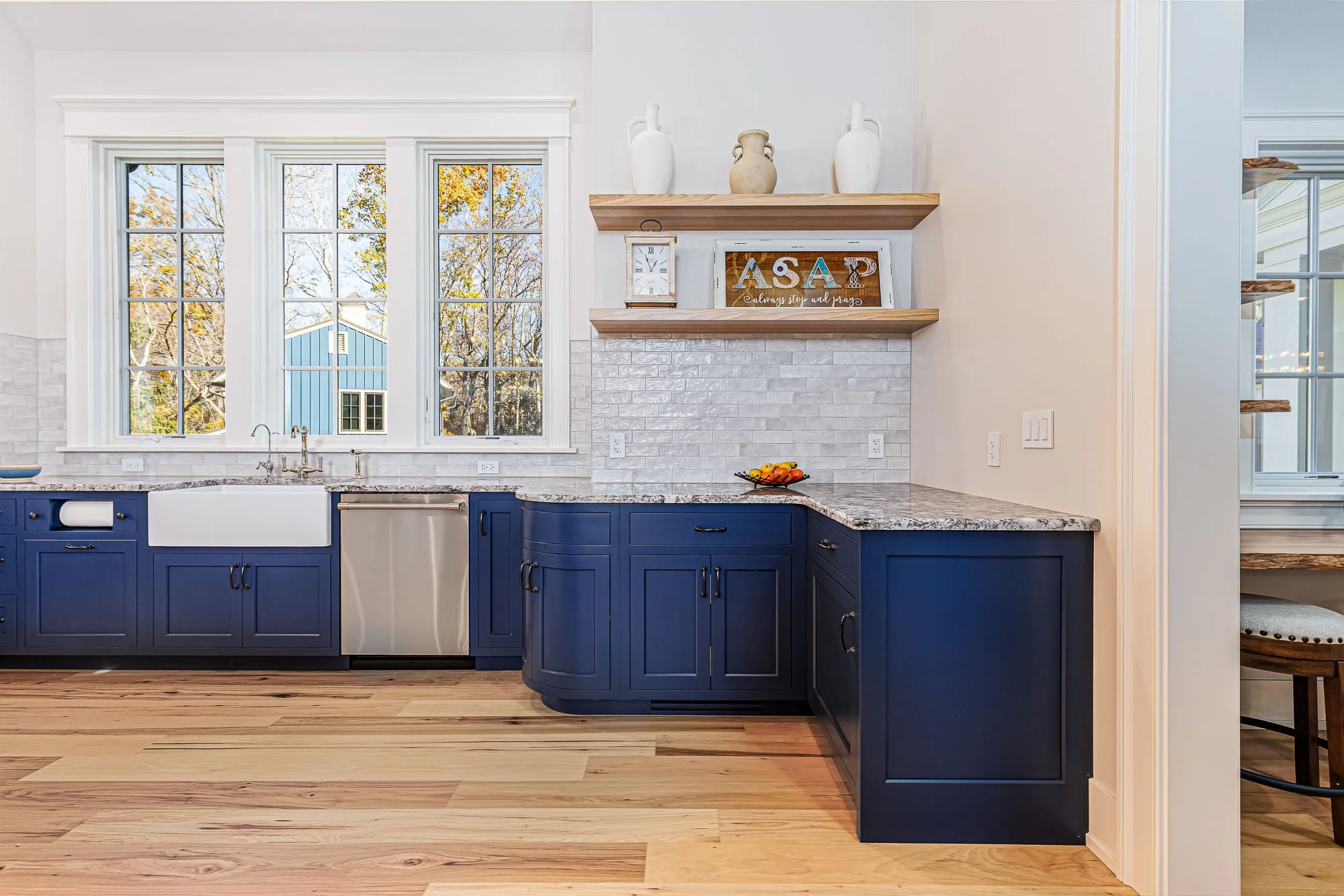 A modern kitchen with blue cabinets, a white farmhouse sink, and a granite countertop. There are three large windows above the sink with a view of trees and a blue building outside. Two wooden shelves hold decorative white and beige vases, with a clock and a sign with the letters 'AΣP' on the middle shelf. The kitchen features light-colored hardwood floors.