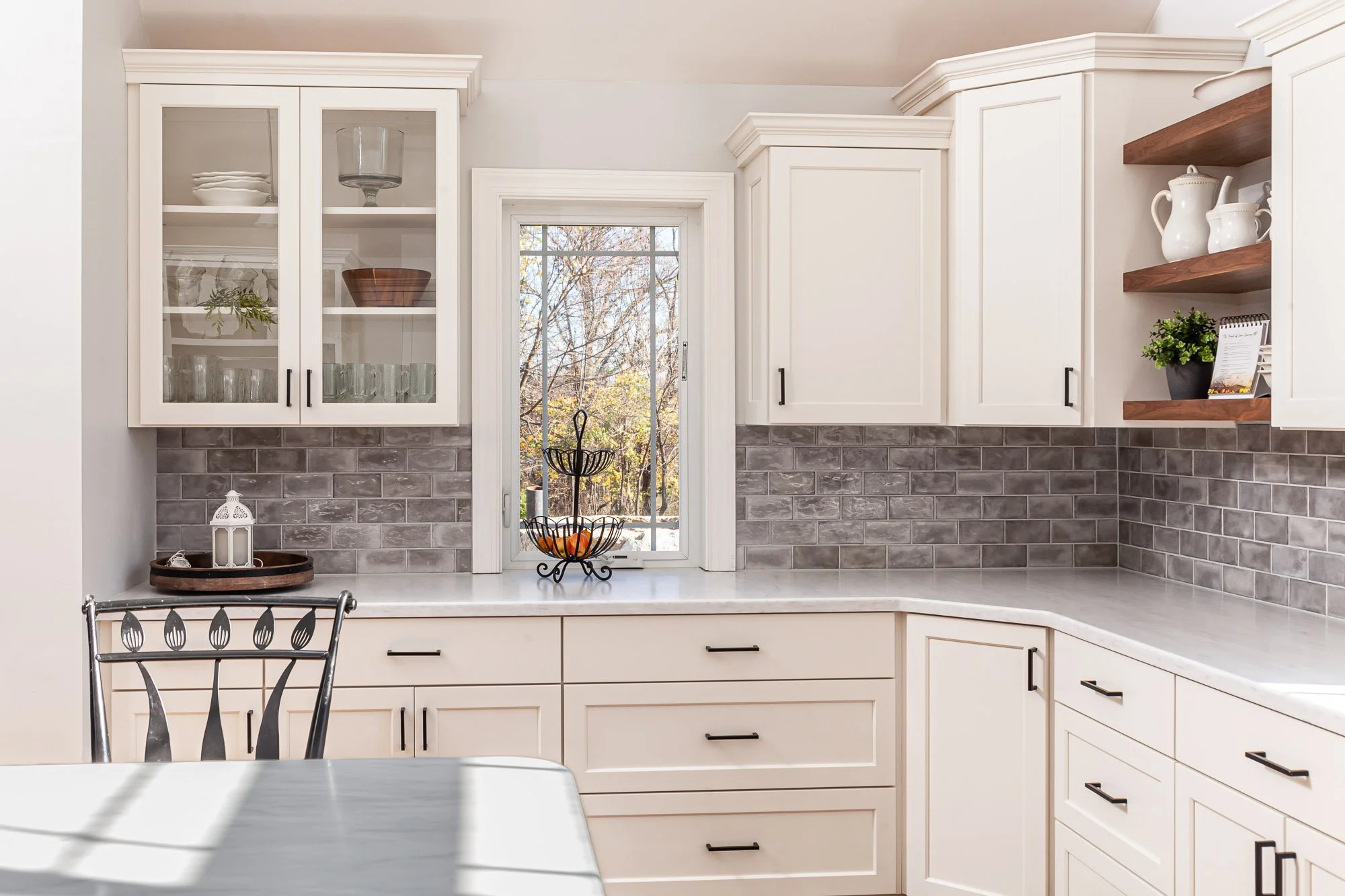 A kitchen with white cabinets, a gray tile backsplash, a window showing trees outside, and a countertop with decorative items including a bowl of oranges and a white lantern.