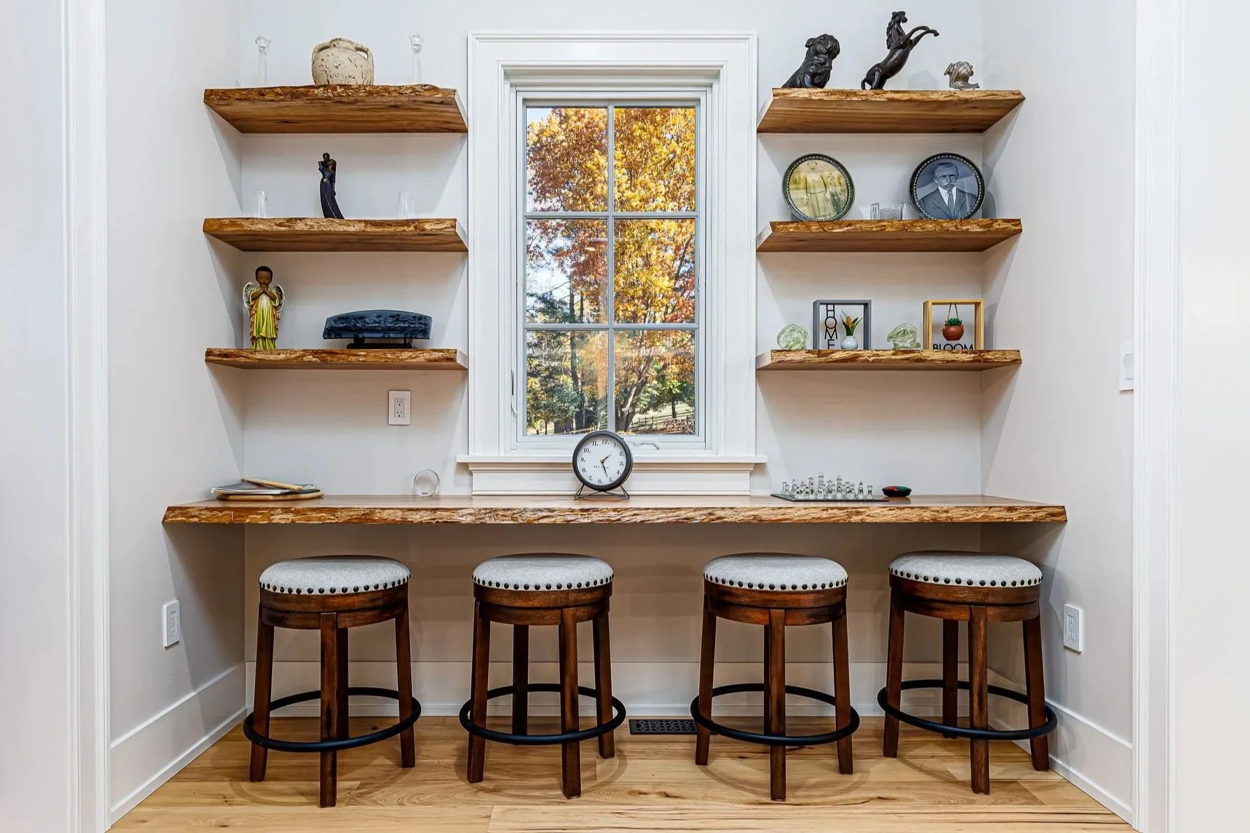 A cozy breakfast bar with four wooden stools with beige cushions, a long wood countertop, a clock and a chess set underneath a window, with wooden shelves above displaying decorative items, art pieces, and a scenic autumn view outside.