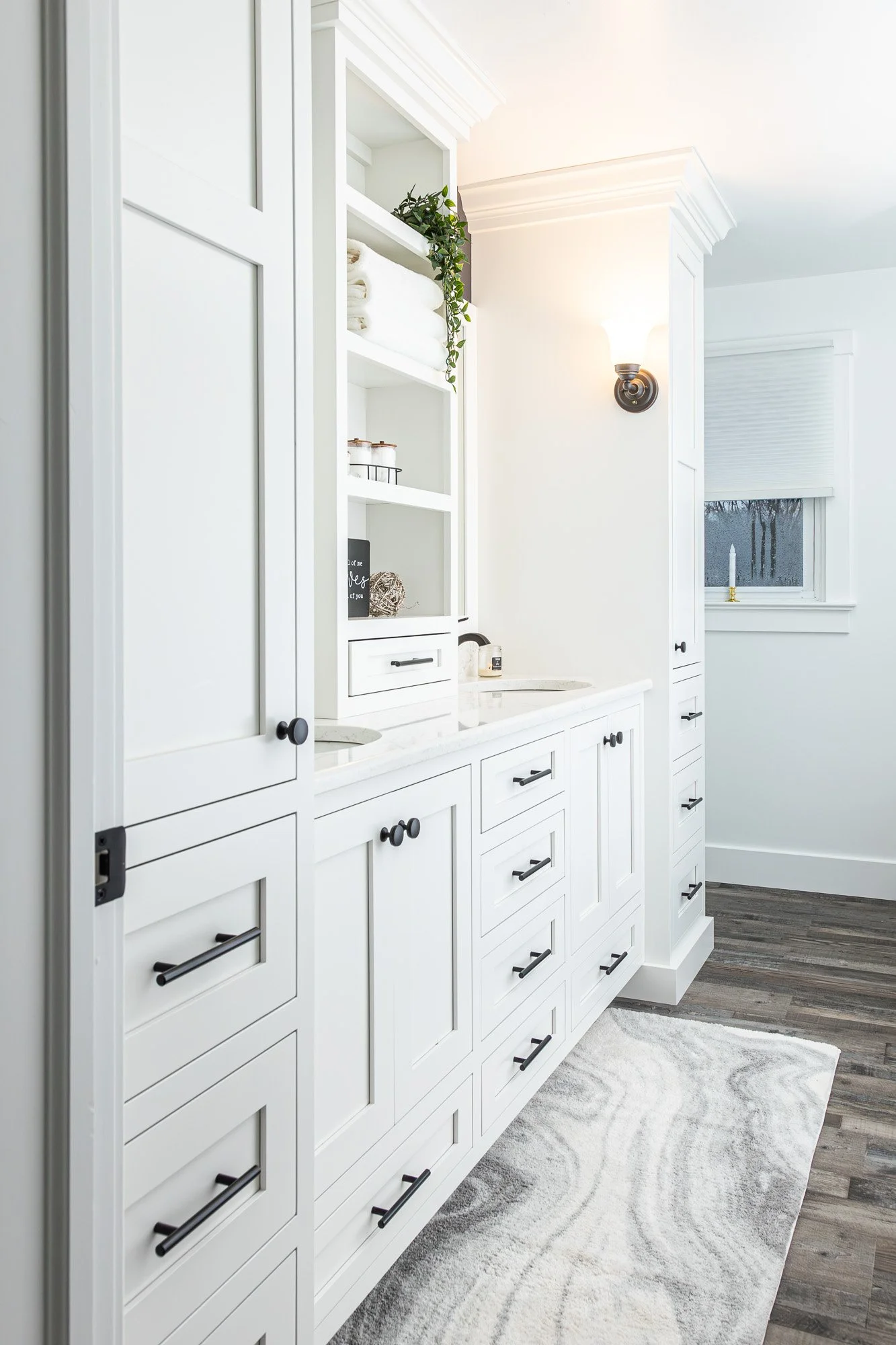 A white kitchen with multiple cabinets, black handles, a window, a sconce light fixture, towels, decorative objects, and a light-colored rug on wooden flooring.