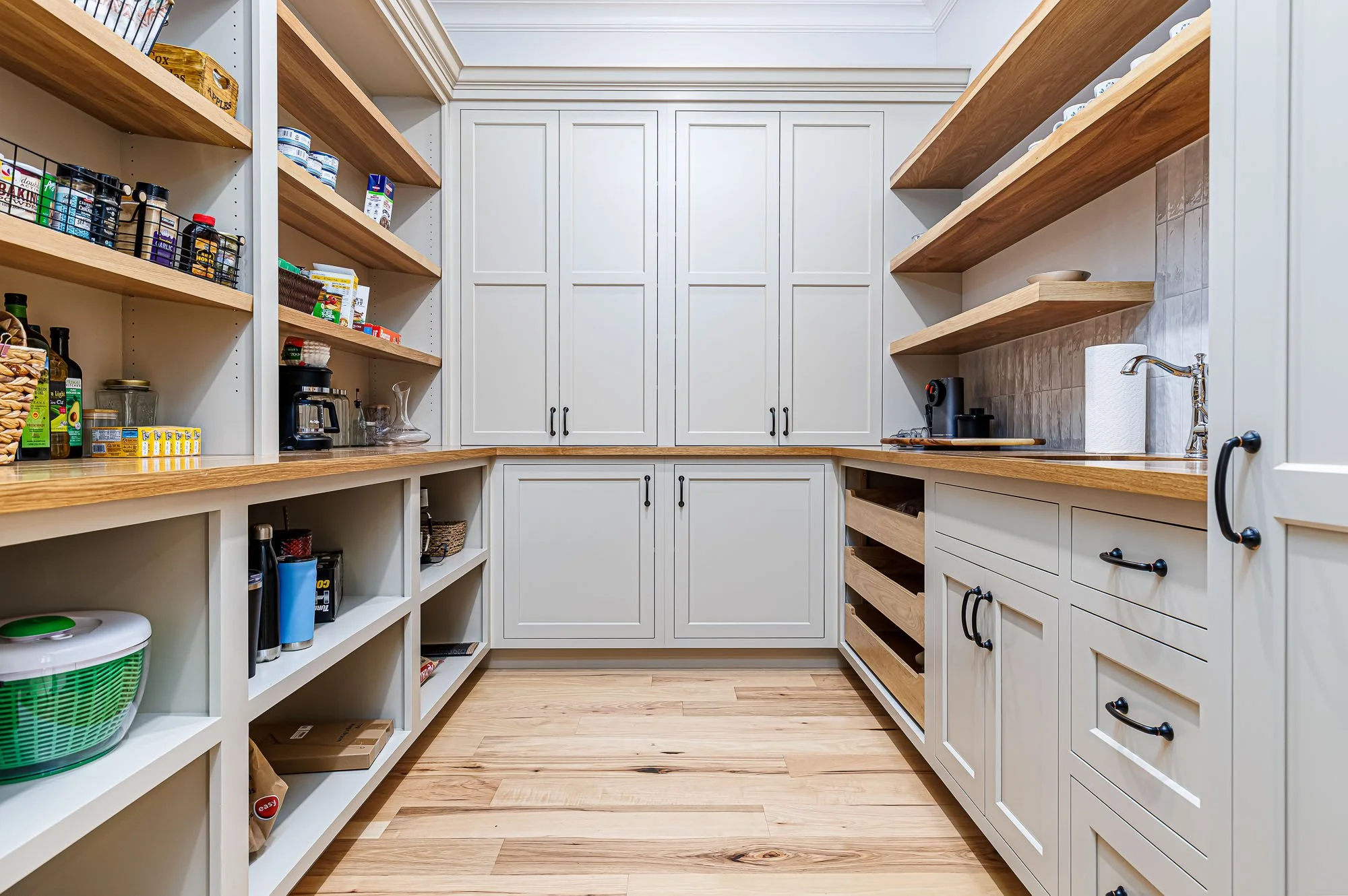 A well-organized walk-in pantry with white cabinets, open wooden shelves, and a wooden countertop. Shelves are stocked with food items, jars, and kitchen supplies, with a coffee machine and a sink visible.