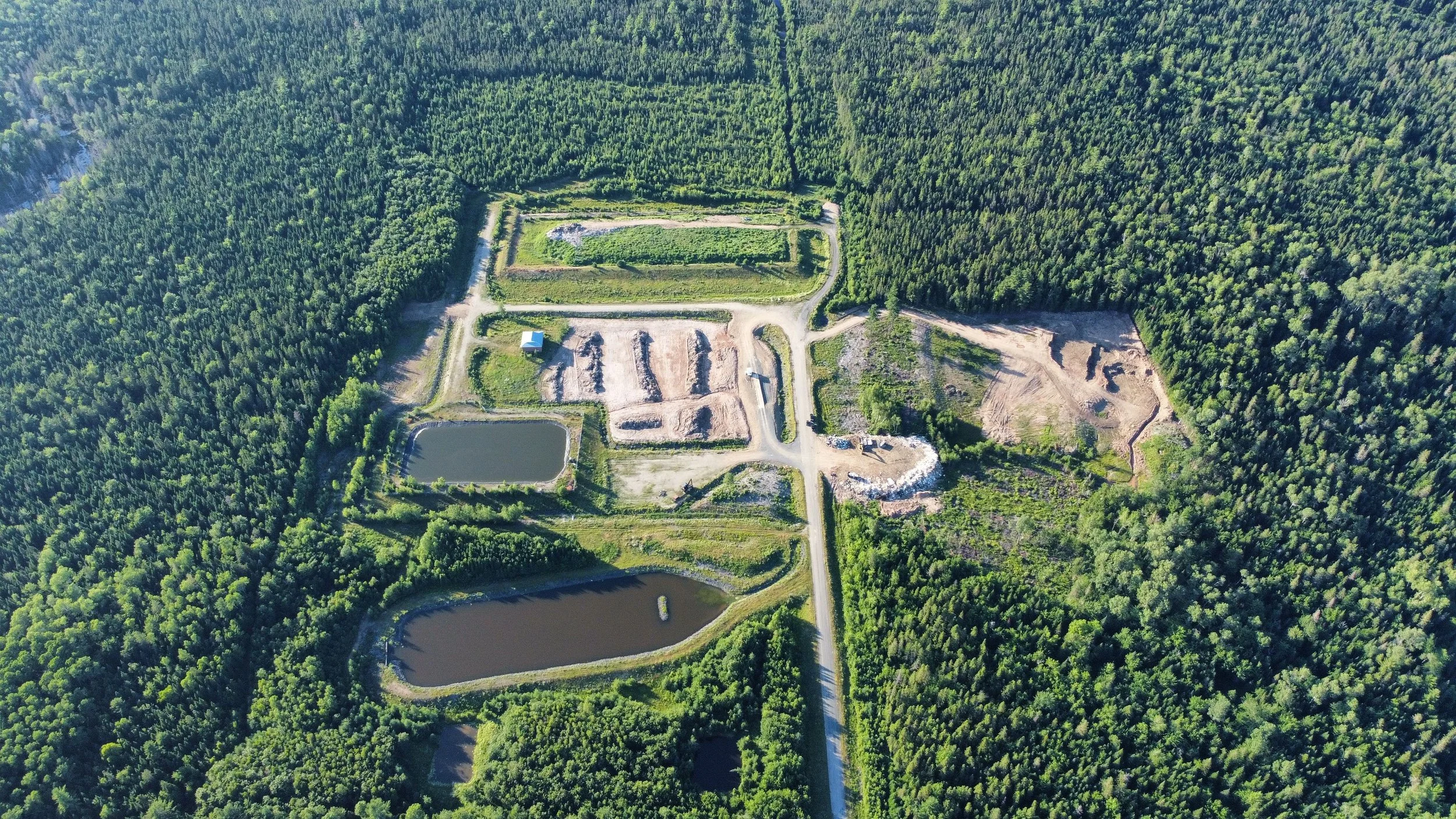 An aerial view of a forested area with a small construction site or excavation area, a pond, and a few small structures, connected by dirt roads.
