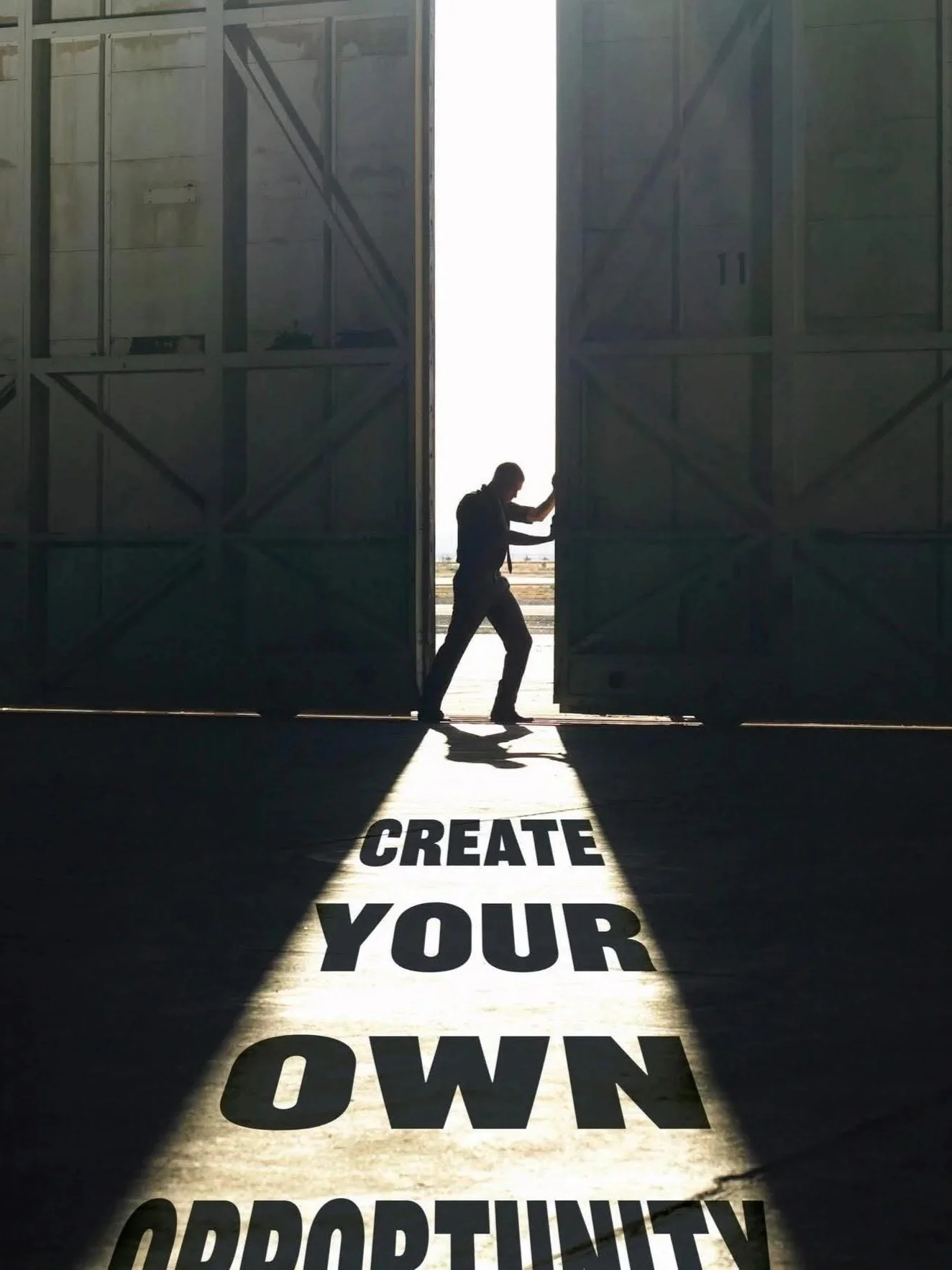 A person walking through large open warehouse doors with sunlight streaming in, casting the phrase "CREATE YOUR OWN OPPORTUNITY" on the floor.