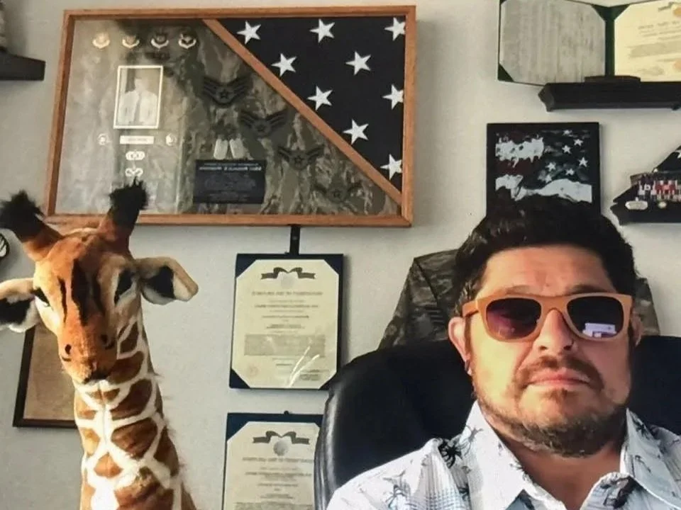 A man with short dark hair, wearing sunglasses and a patterned shirt, taking a selfie in an office. To his left, a giraffe plush toy is standing on its head. Behind them on the wall are military or patriotic decorations, including framed certificates, a display case with medals, and a flag with stars and stripes.