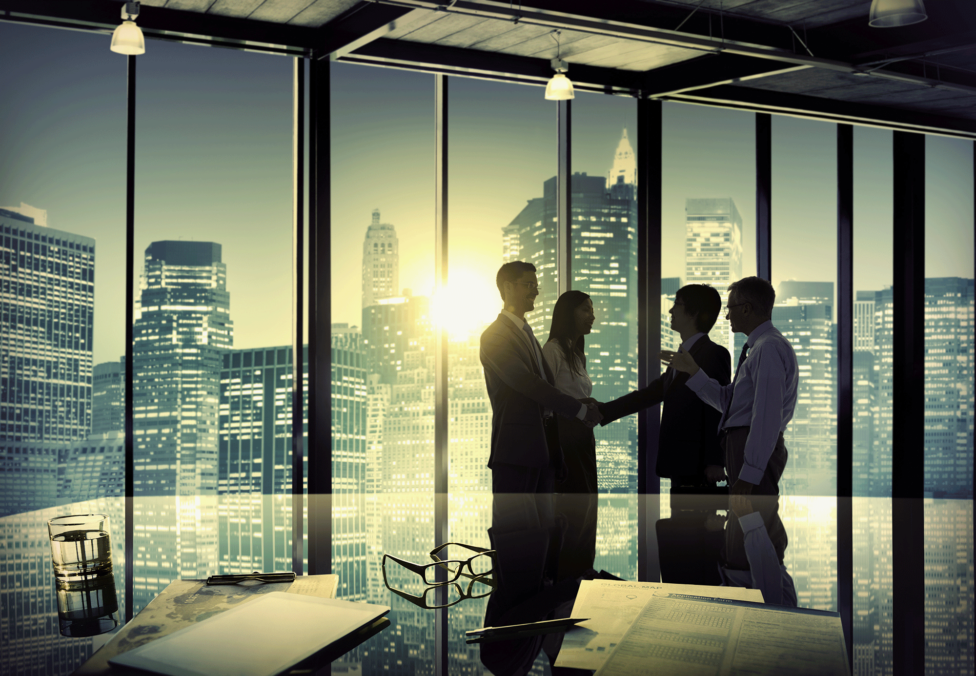 Silhouettes of four businesspeople shaking hands in a modern office with floor-to-ceiling windows showing a city skyline during sunset.