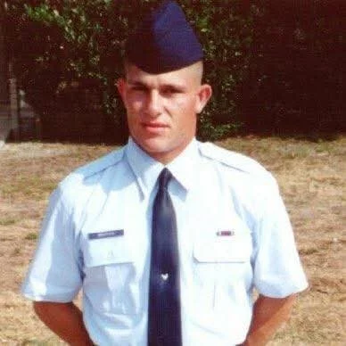 Young man in a military uniform with a blue cap and white shirt, standing outdoors with trees and grass in the background.