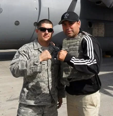 Two men, one in military uniform and sunglasses, and the other in casual clothing with a cap, are posing with fists raised in front of an airplane.