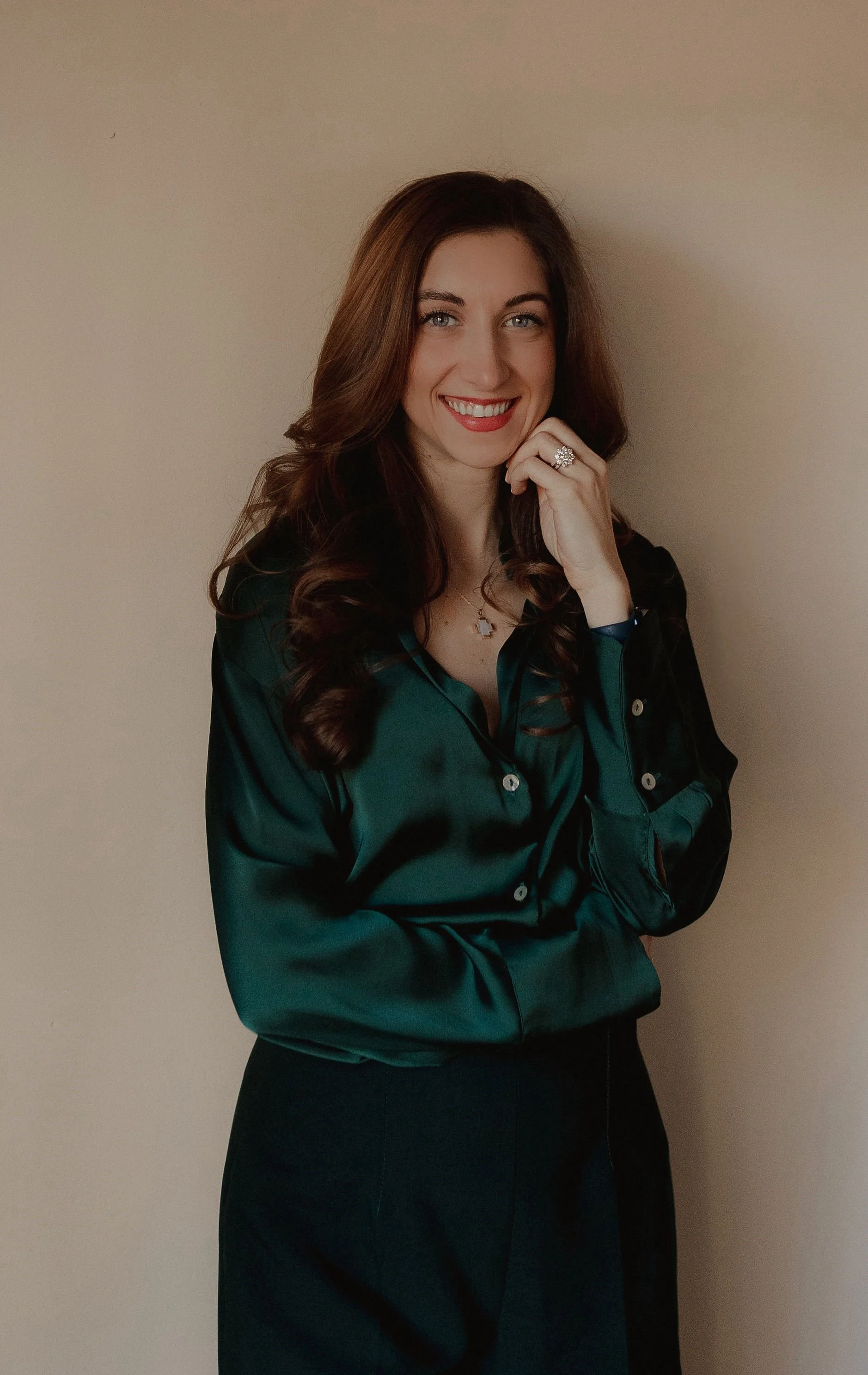A woman with long brown hair, blue eyes, and fair skin is smiling. She is wearing a dark green satin blouse, a ring, and a necklace, and is standing against a plain beige wall, with her left hand resting near her chin.