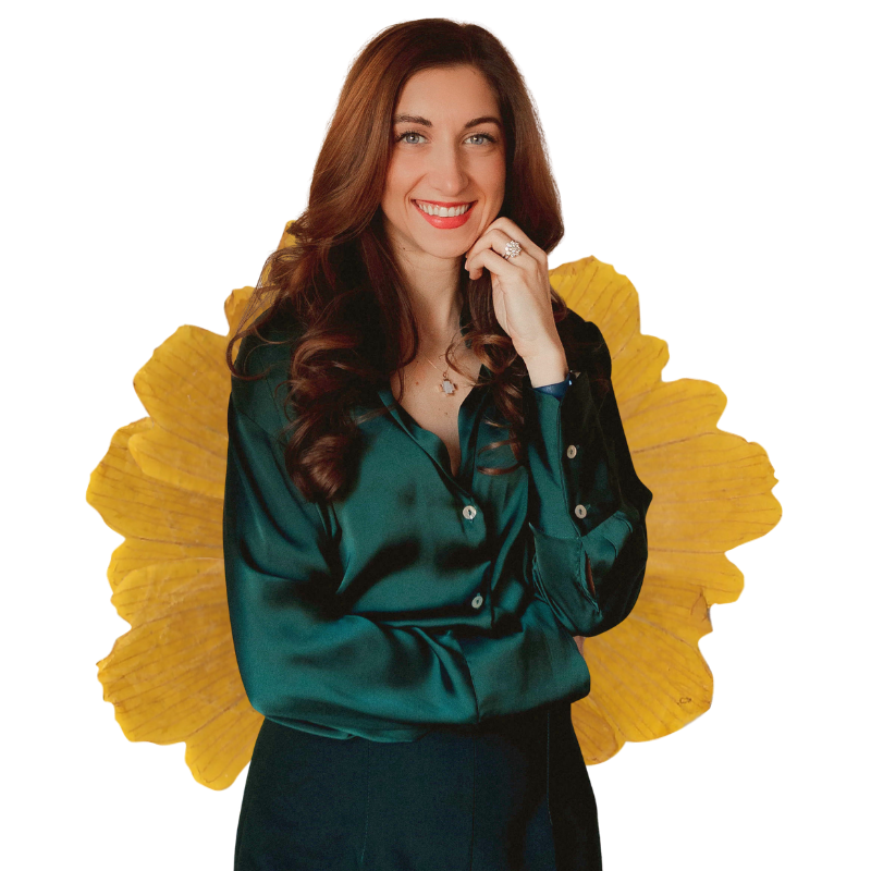 A woman with long brown hair, wearing a dark green satin blouse, smiling, with a large yellow flower behind her.