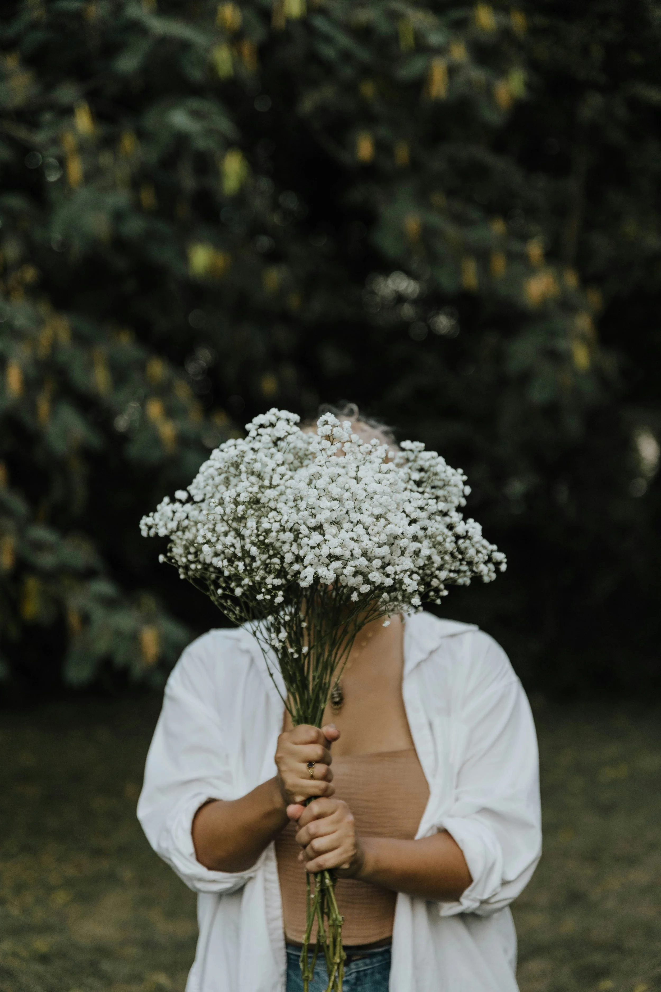 A person standing outdoors holding a bouquet of white flowers that cover their face, with a background of dark green foliage.