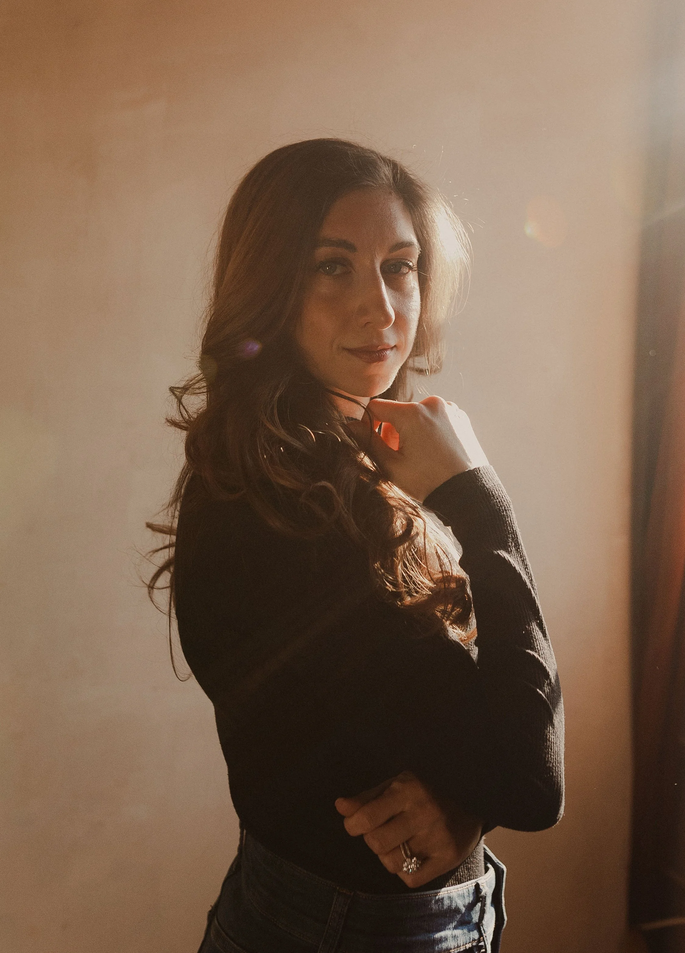 A woman with long, wavy brown hair and light makeup, wearing a black long-sleeve top and dark jeans, stands against a softly lit, neutral background with sunlight creating a warm glow around her.