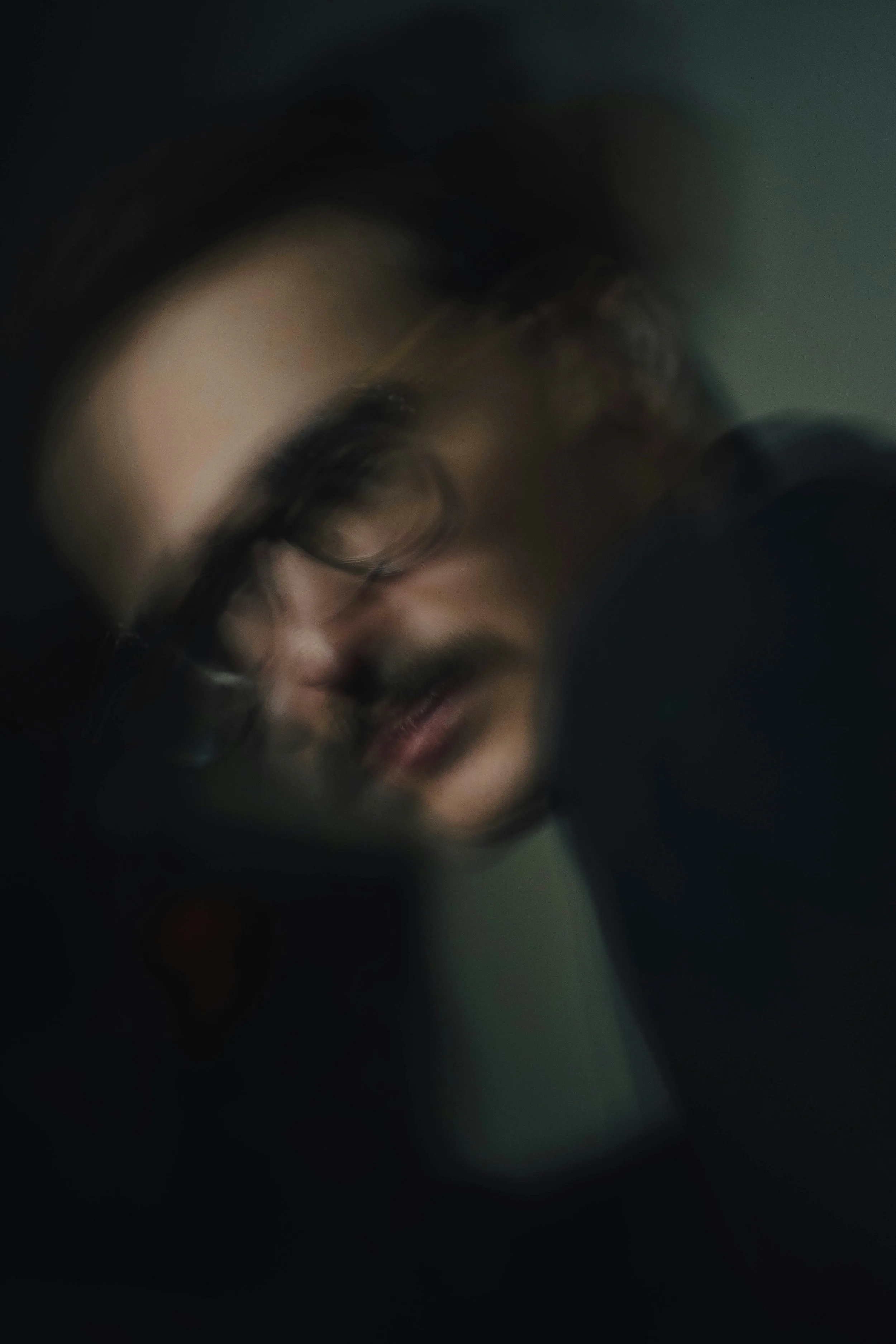 Close-up of a person's face wearing glasses, with a shadowy background, and the face slightly tilted downward.