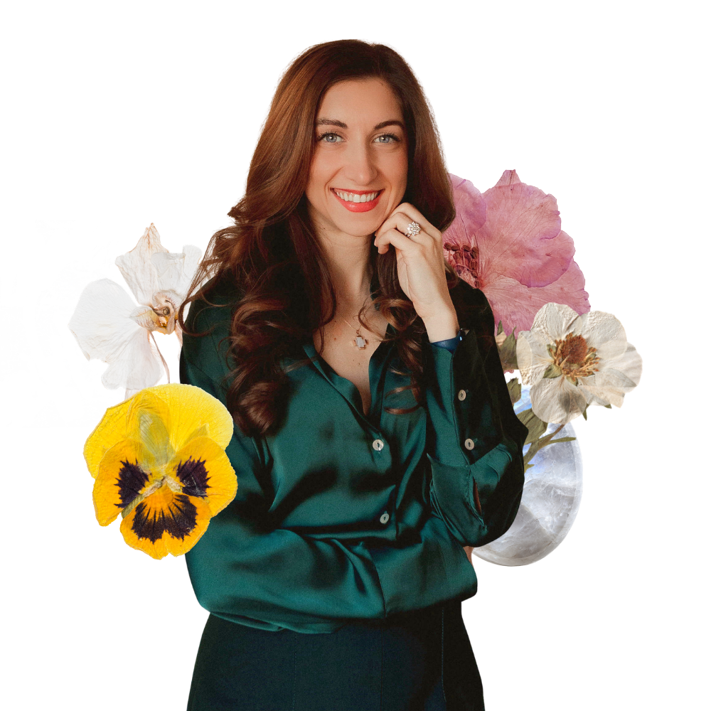 A woman with long brown hair and blue eyes, smiling and wearing a dark green satin blouse, standing in front of a background decorated with colorful flowers.
