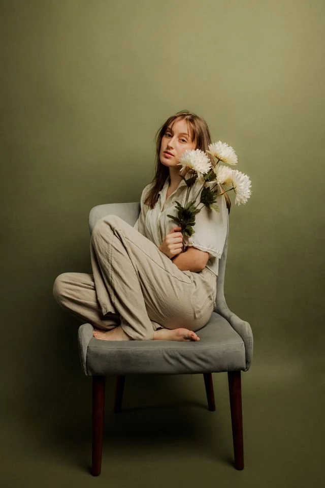 a woman sits in a tan chair in a tan outfit, blank expression on her face, her face partially covered by flowers