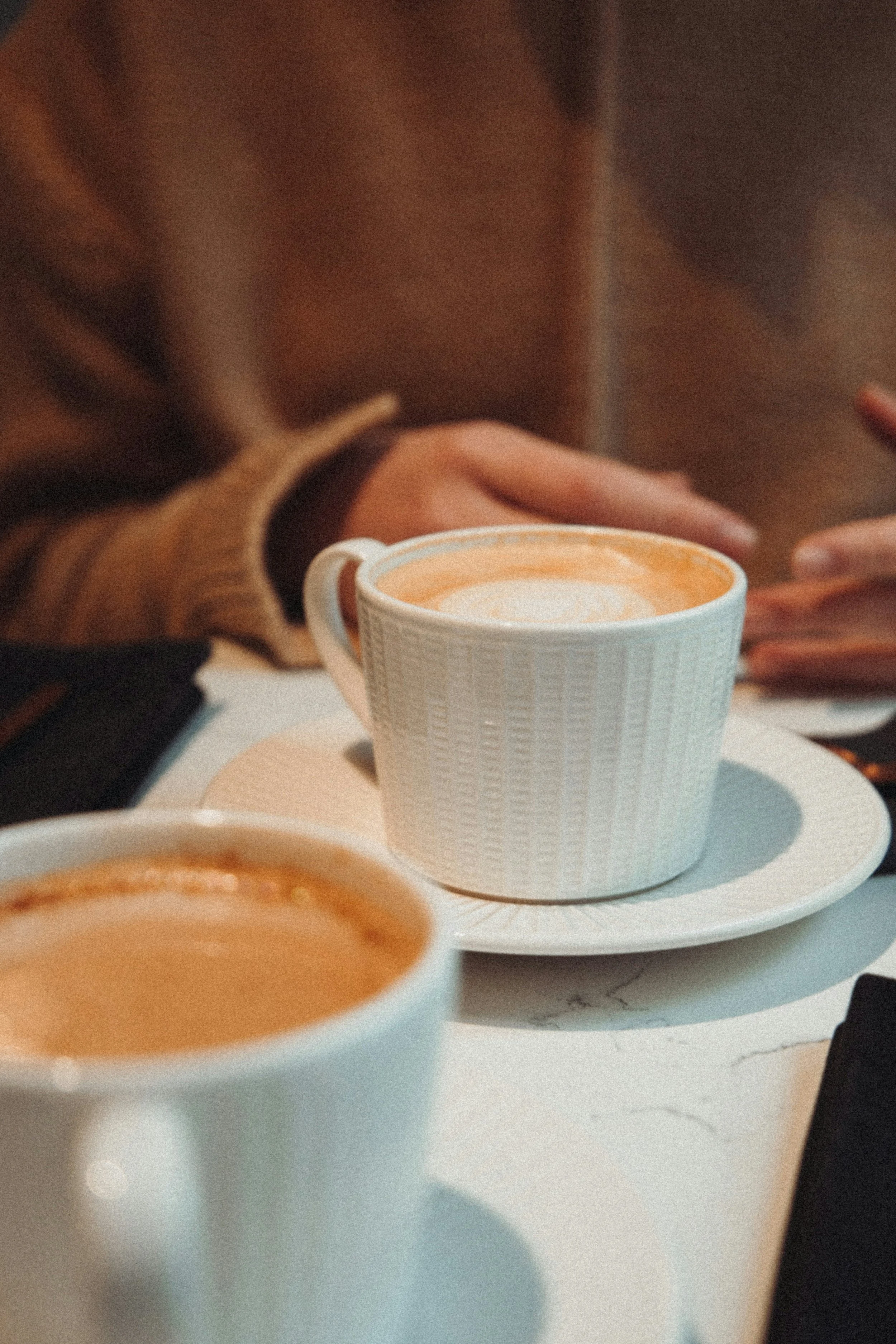 Close-up of two cups of coffee on a white table, with a person in a brown hoodie sitting in the background