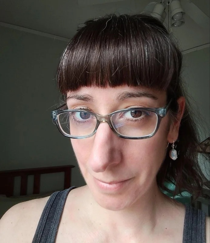 A woman with brown hair and bangs wearing blue glasses and white-and-blue earrings.