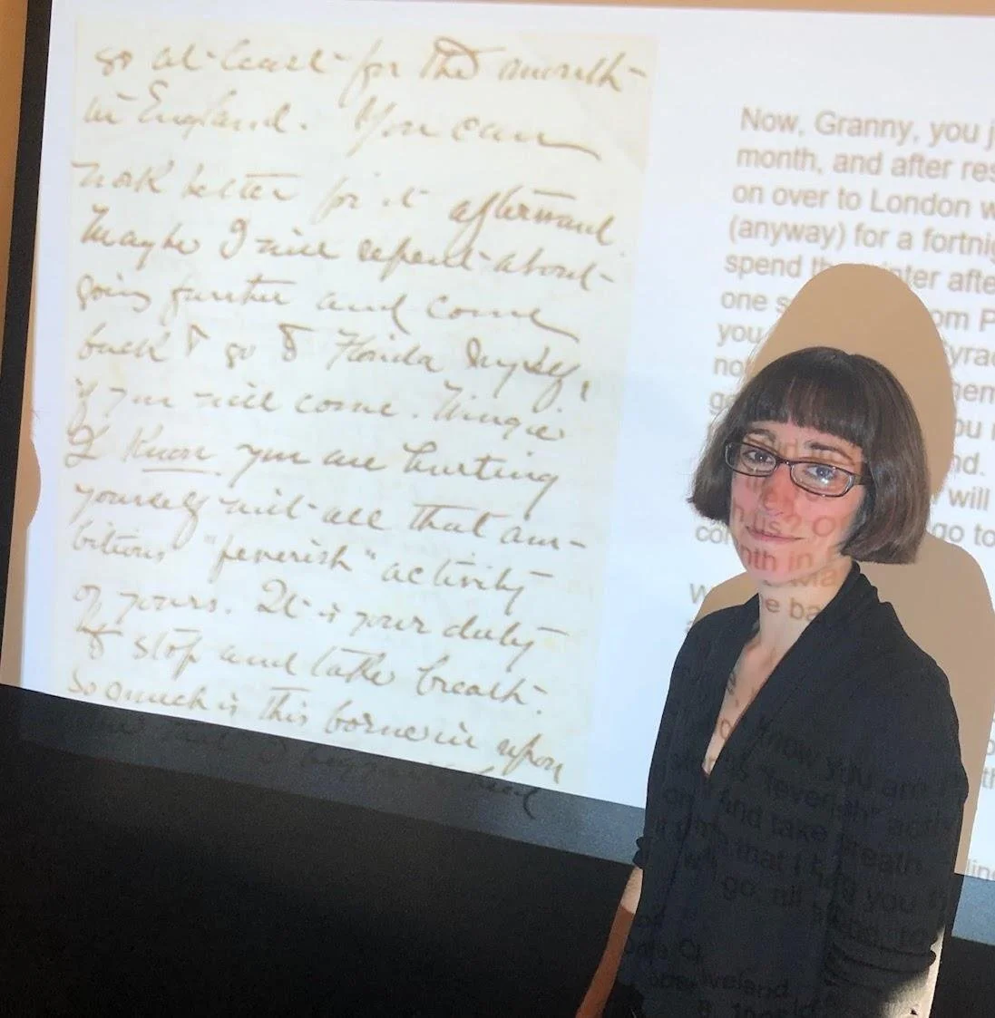 A woman with brown hair in a bob, glasses, and a black sweater standing in front of the image of a handwritten letter projected onto a screen.