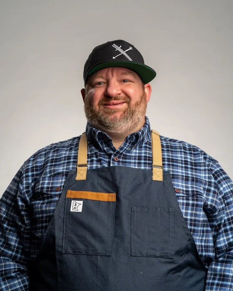 A man wearing a black baseball cap with a white archer and arrow design, a blue plaid shirt, and a dark apron, smiling in front of a plain background.