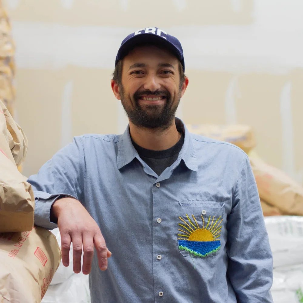 A man with a beard and mustache smiling, wearing a blue baseball cap and a light blue button-up shirt with a sun and wave embroidered patch on the pocket, standing in a store aisle with bags of rice or flour behind him.
