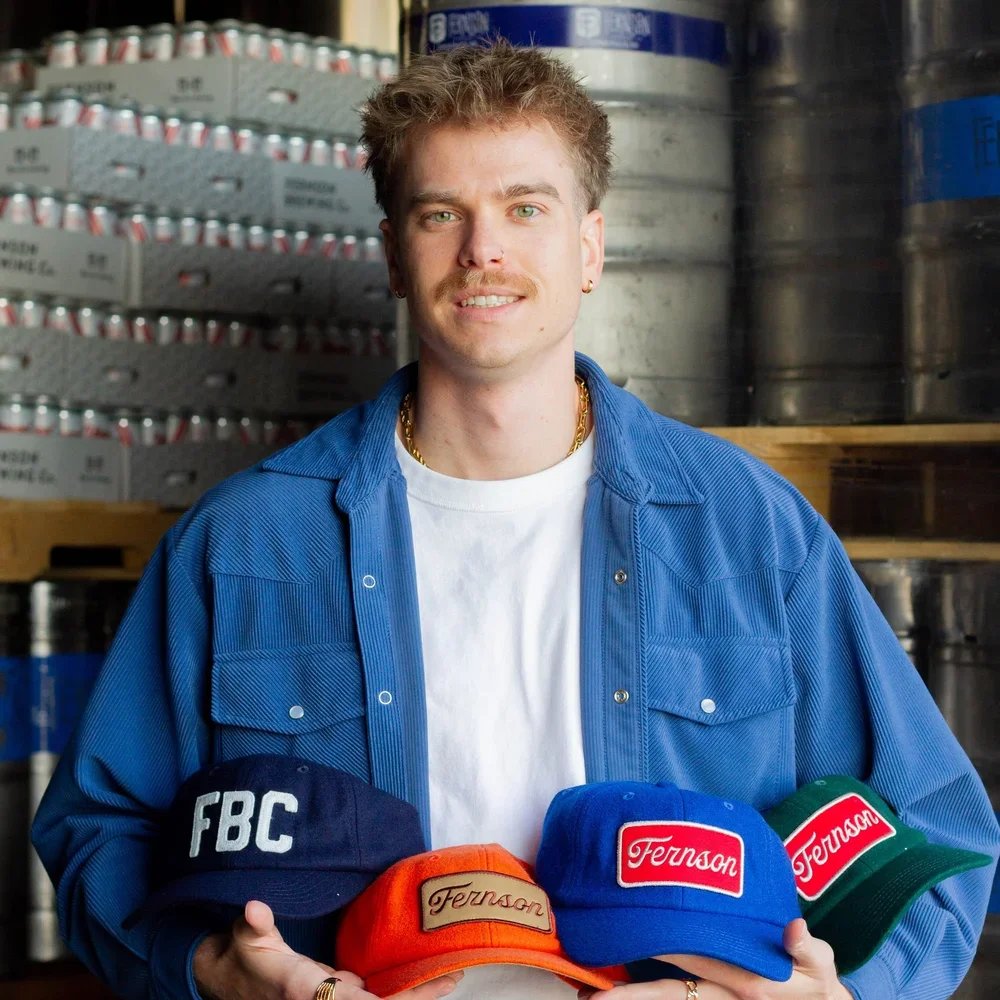 A young man with curly hair, a mustache, and earrings, smiling and holding four colorful hats with brand logos in a warehouse of canned and bottled beverages.