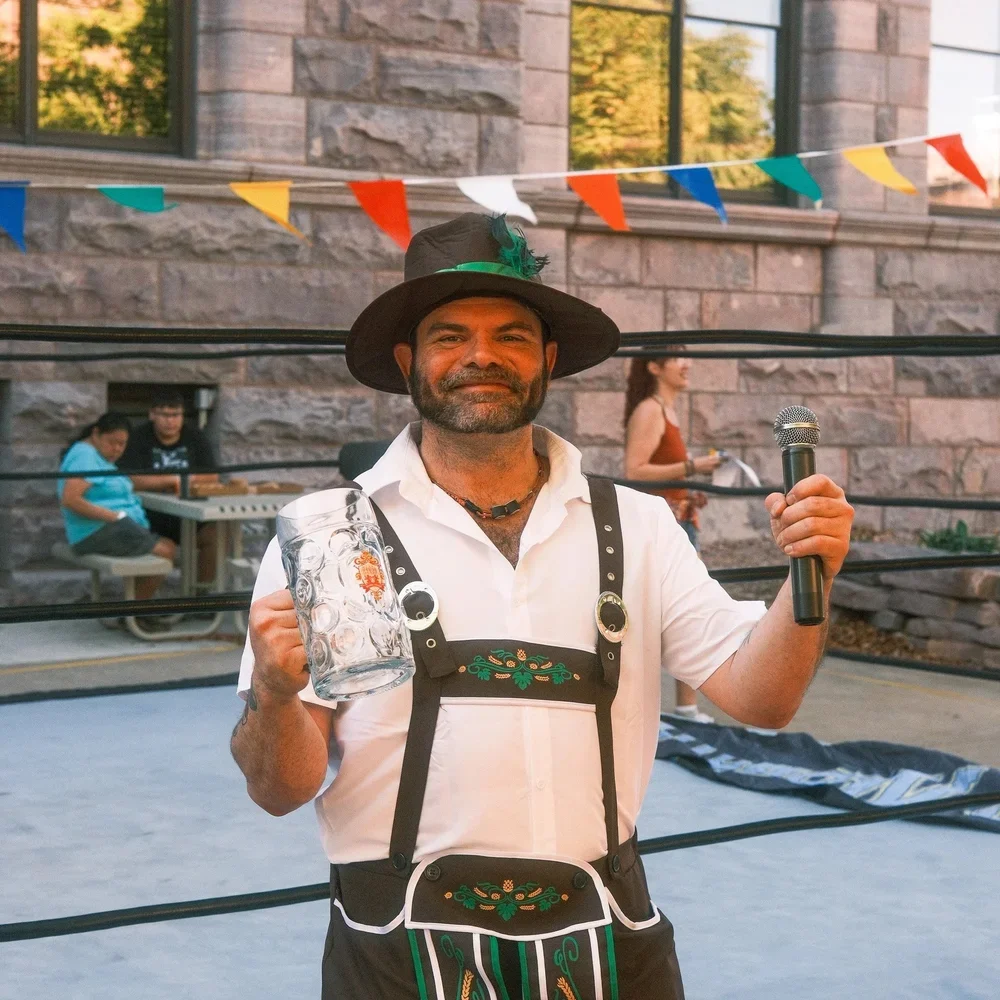 A man dressed in traditional Bavarian attire, holding a large beer stein in one hand and a microphone in the other, standing inside a ring with colorful decorations.