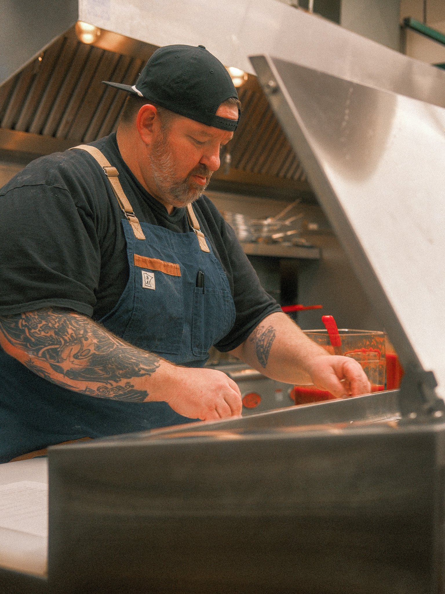 A man wearing a black cap backwards, a black shirt, and a blue apron with leather straps is preparing food in a commercial kitchen. He has tattoos on his arms and is focused on his work with a glass of red sauce or ketchup nearby.
