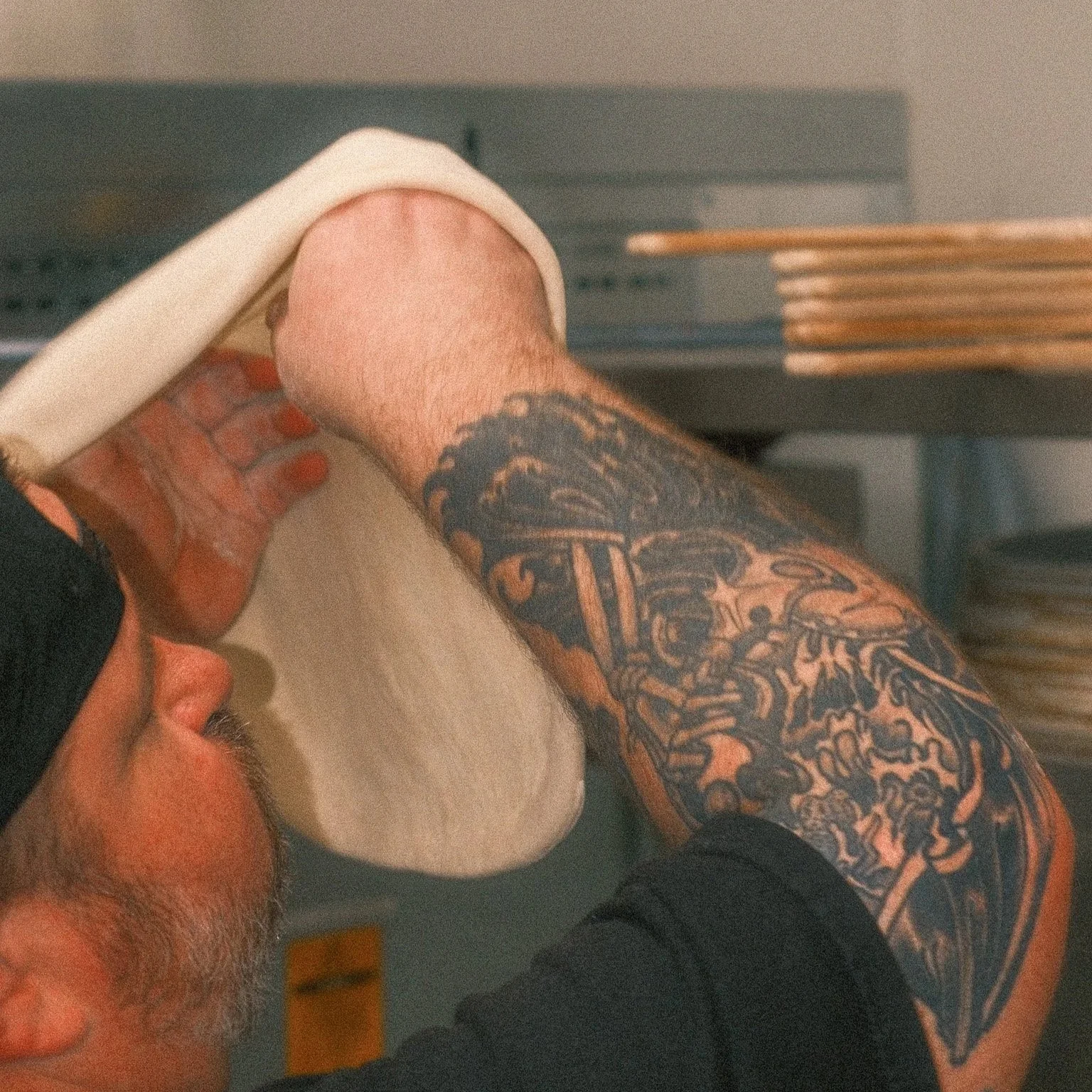 A man with tattoos on his arm and shoulder, lying on his back as another person shaves his face with a razor.