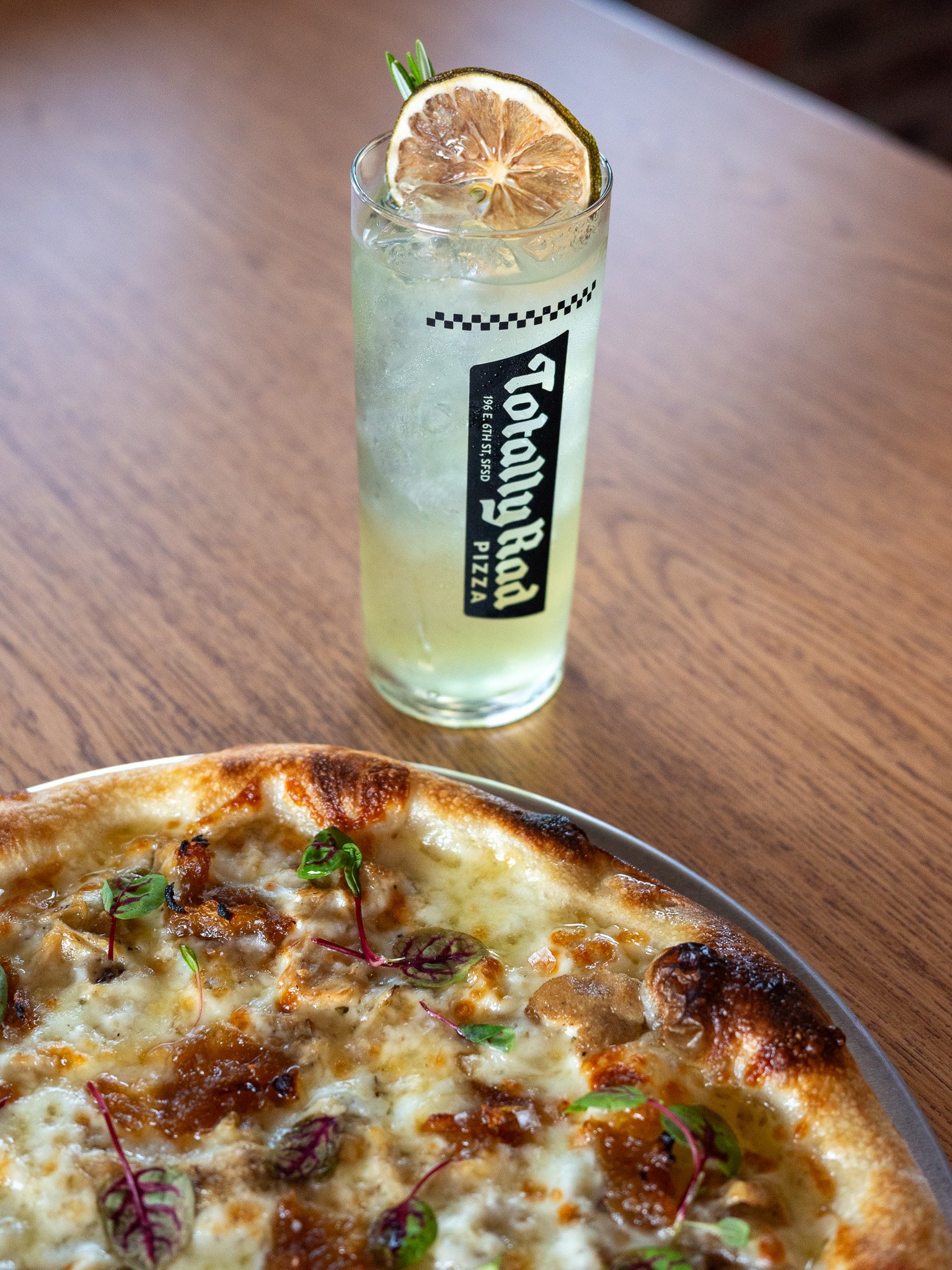 Close-up of a pizza with cheese, toppings, and microgreens on a wooden table, with a glass of lemon soda garnished with lemon slices and a sprig of herbs in the background.