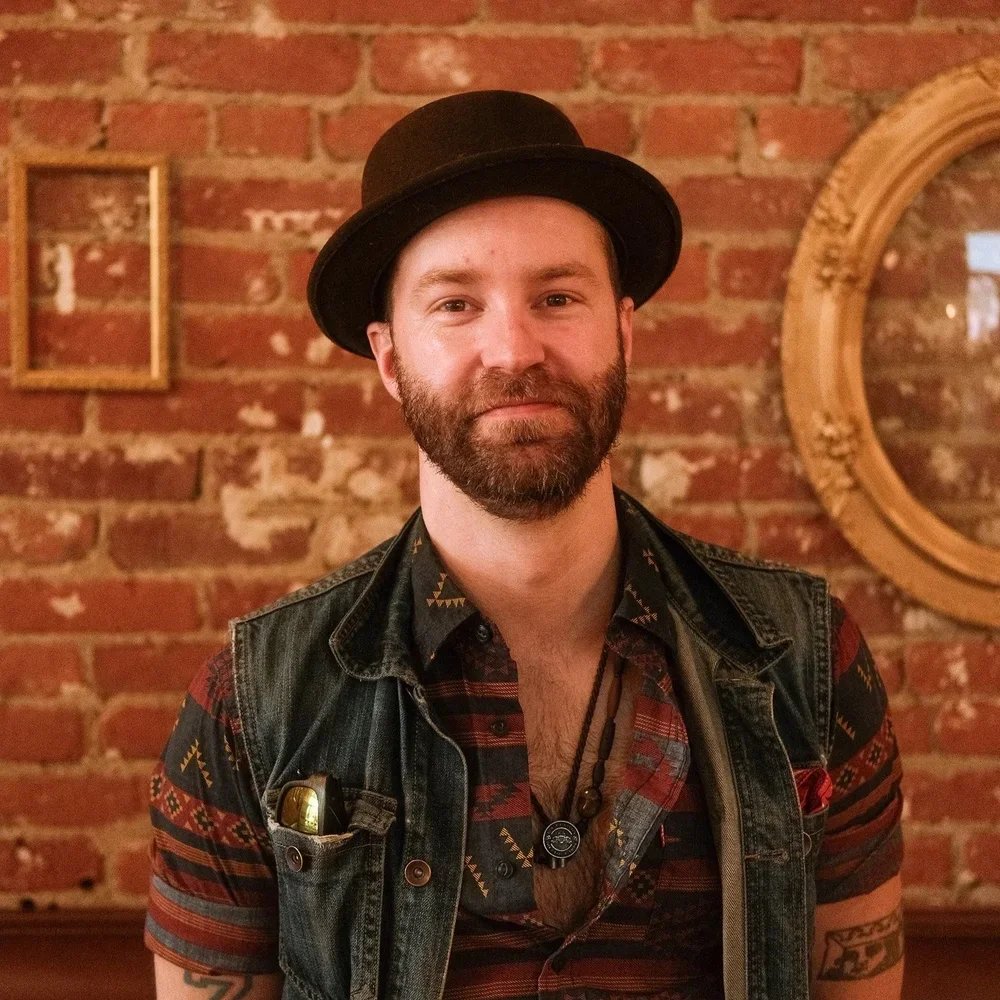 A man with a beard wearing a black hat and a patterned shirt, standing against a brick wall with picture frames and a round mirror.