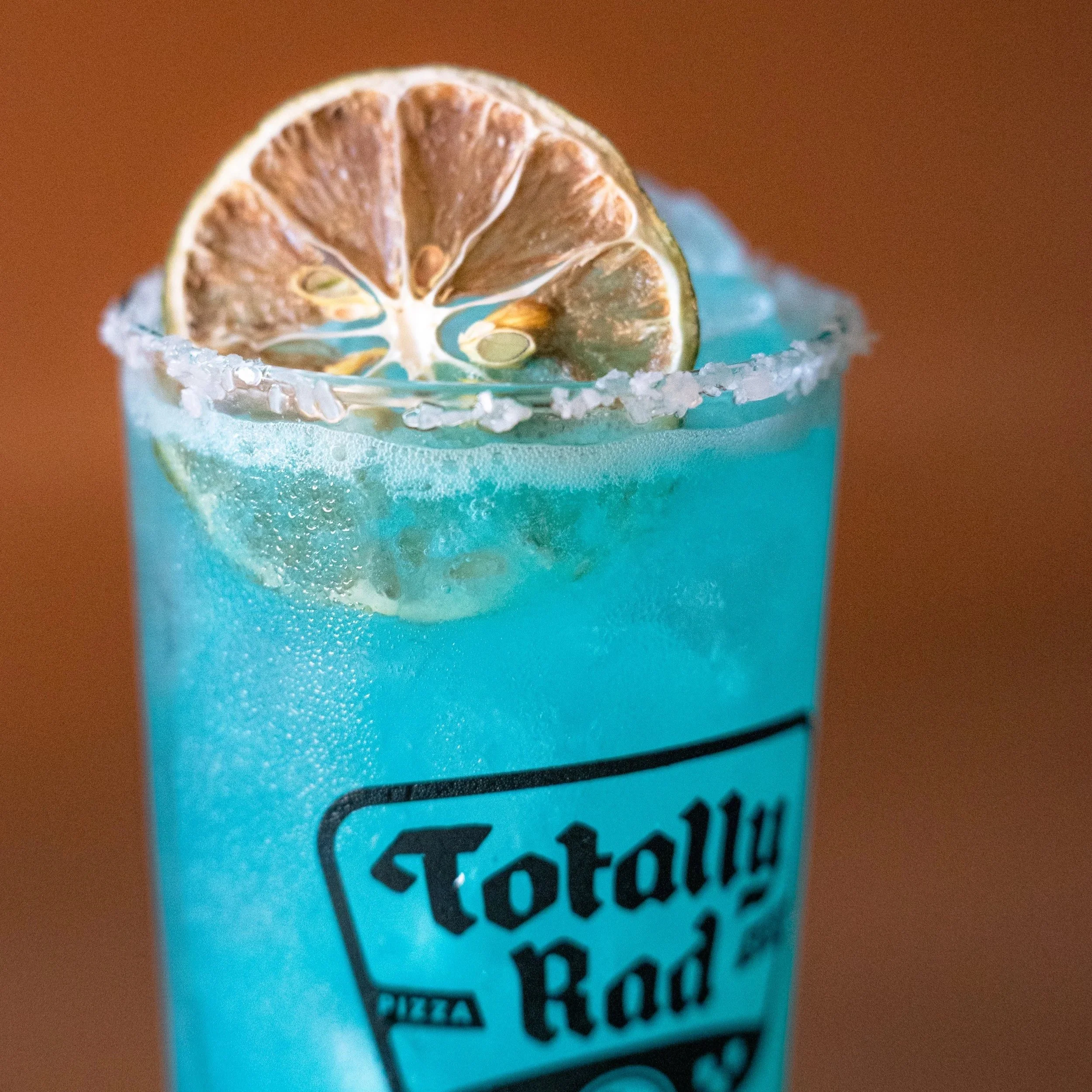 Close-up of a blue cocktail in a glass rimmed with salt, topped with a dried lime slice, with part of a menu or sign below reading 'something'.}
