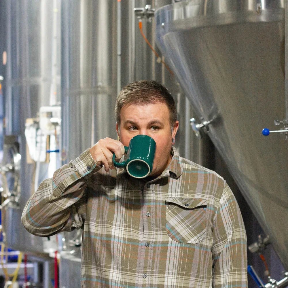 A man with short hair wearing a plaid shirt, drinking from a green mug in an industrial setting with large metal tanks.