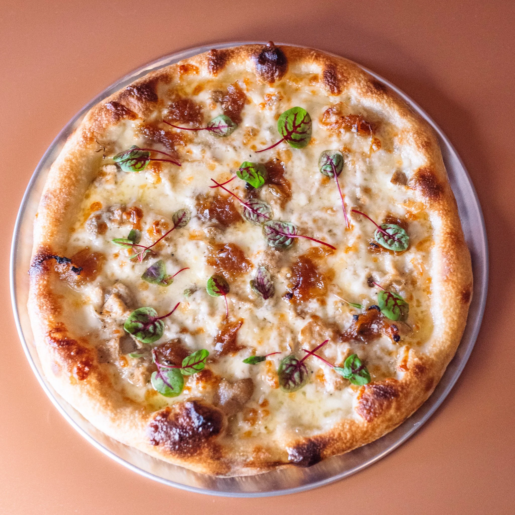 A pizza with a golden-brown crust topped with melted cheese, small greens, caramelized onions, and possibly bacon or ham, served on a silver tray.