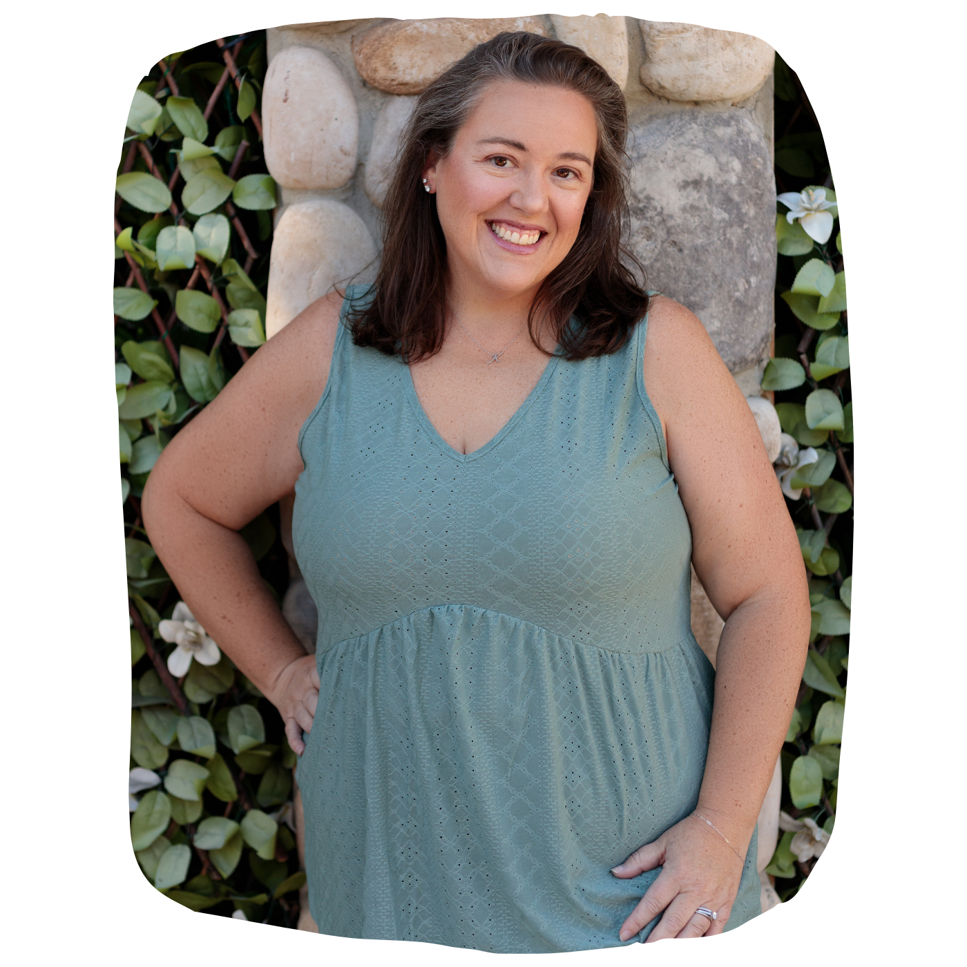 A woman with shoulder-length brown hair, smiling, wearing a sleeveless blue dress, standing outdoors against a stone wall and green leafy background.