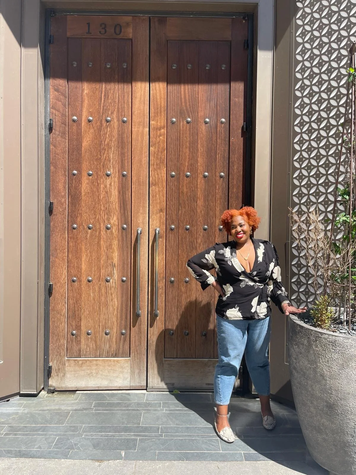 A woman with curly orange hair wearing a black and white floral top and blue jeans, standing in front of a large wooden door with the number 130 above it, smiling outdoors.