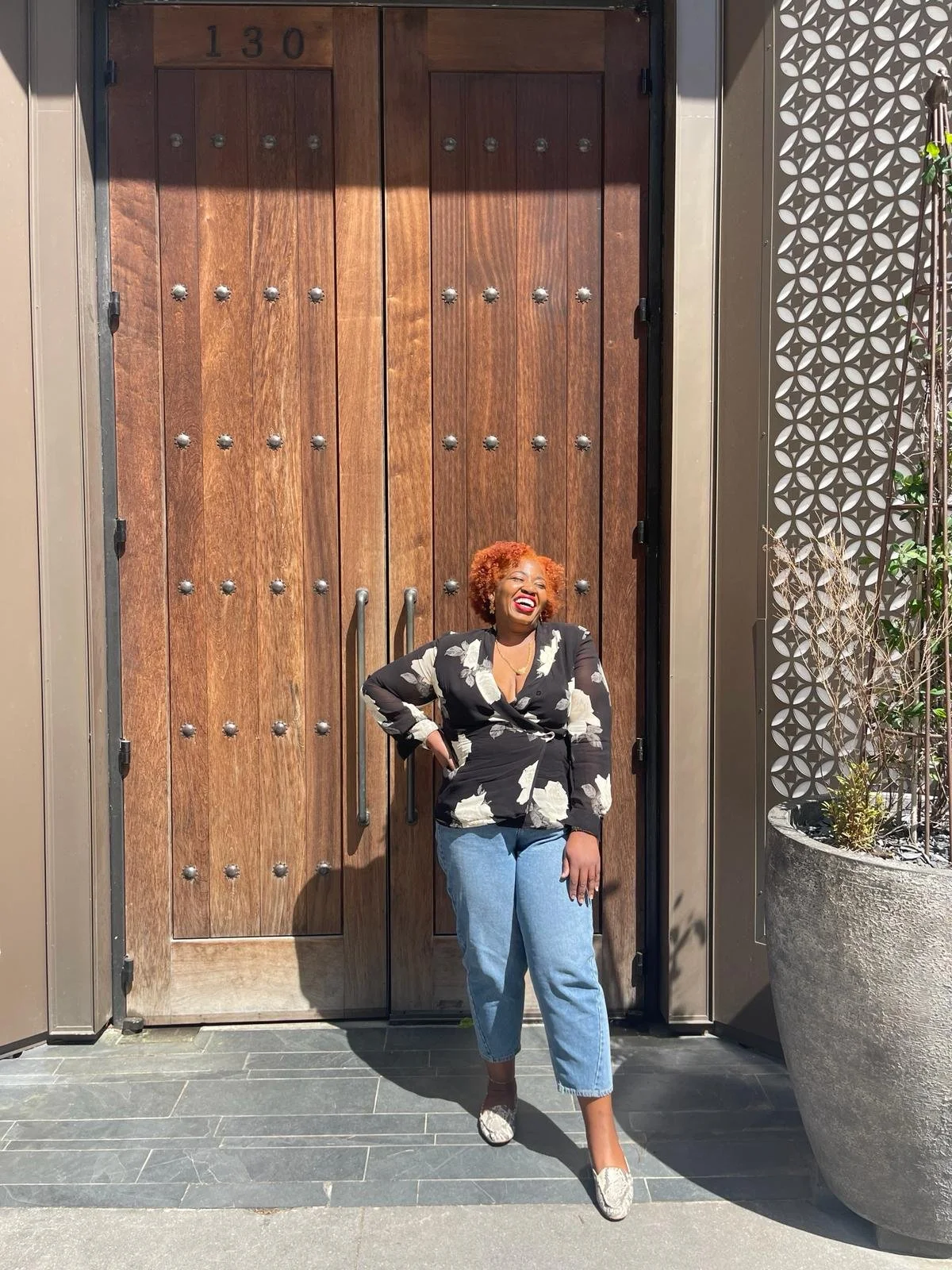 Woman with curly red hair laughing and smiling, standing in front of a large wooden door, wearing a black floral blazer, light blue jeans, and floral shoes, with sunlight shining on her face.
