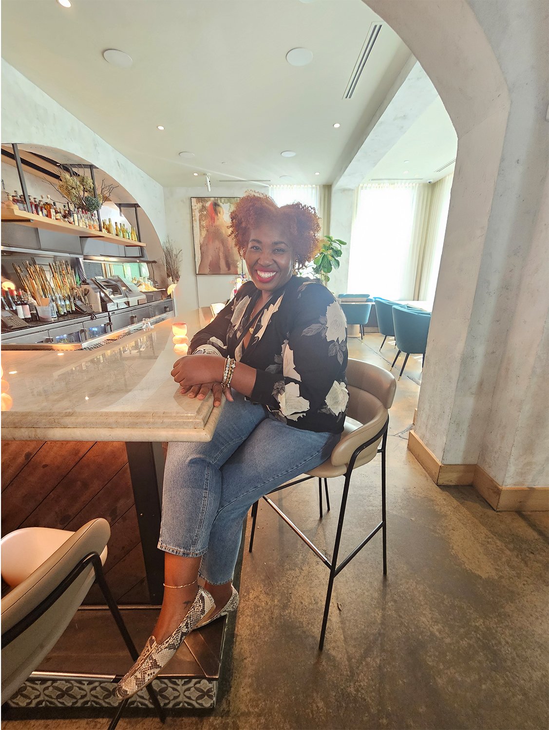 A woman with curly hair and a big smile sitting at a bar counter in a modern, well-lit restaurant or cafe, with a background of bar shelves, artwork, and seating area.