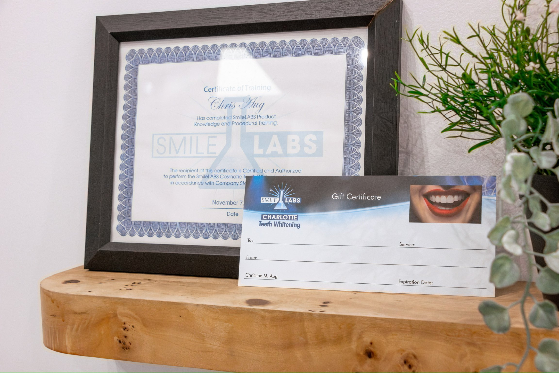 A framed certificate of training from SmileLABS, partially obscured by a flower arrangement on the right, with a separate gift certificate for teeth whitening placed in front of it and a wooden surface underneath.