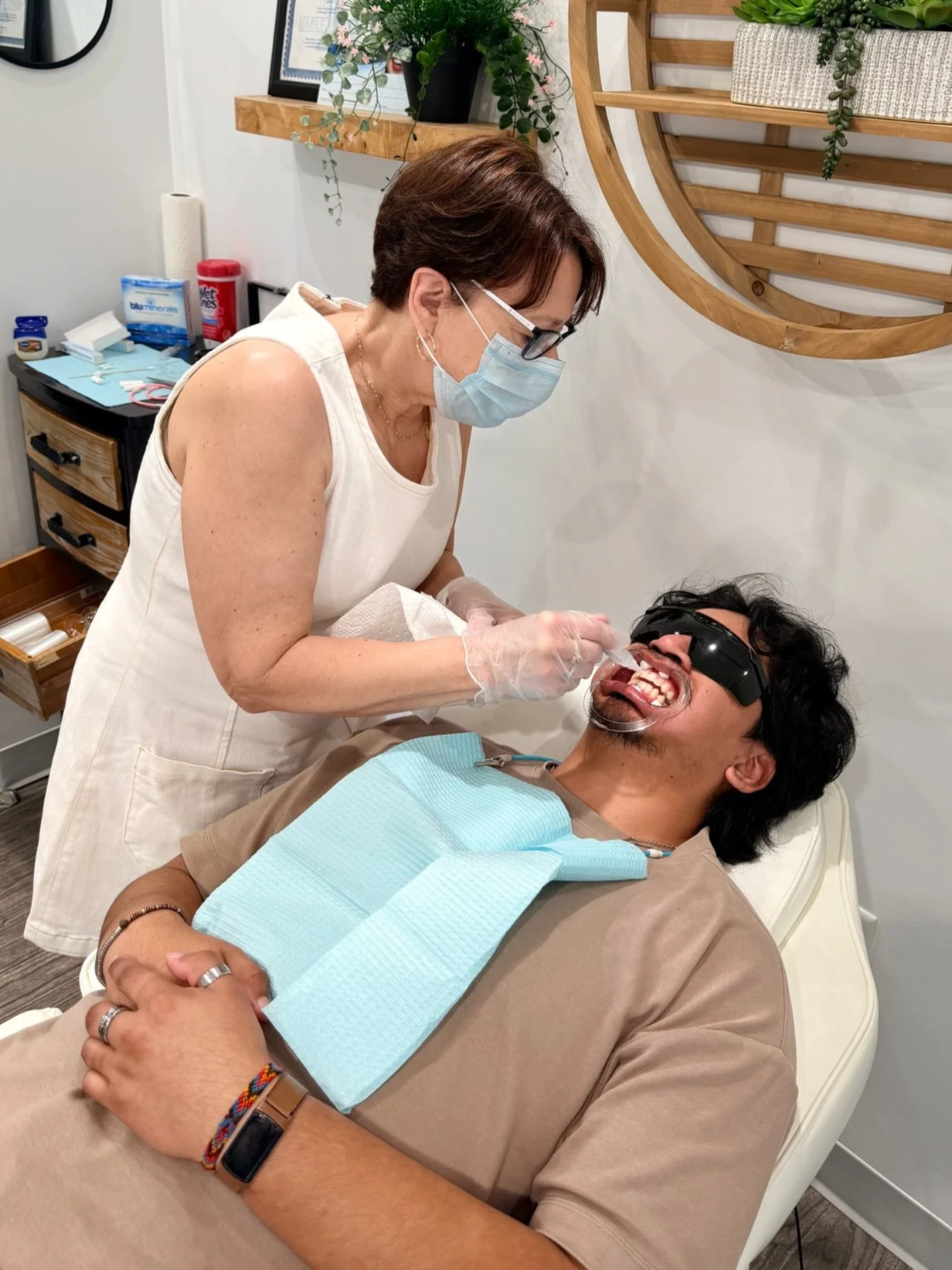 A dental professional (Chris) wearing a mask, gloves, and glasses is examining and cleaning the teeth of a patient reclining in a dental chair. The patient is wearing sunglasses and has a dental bib on.
