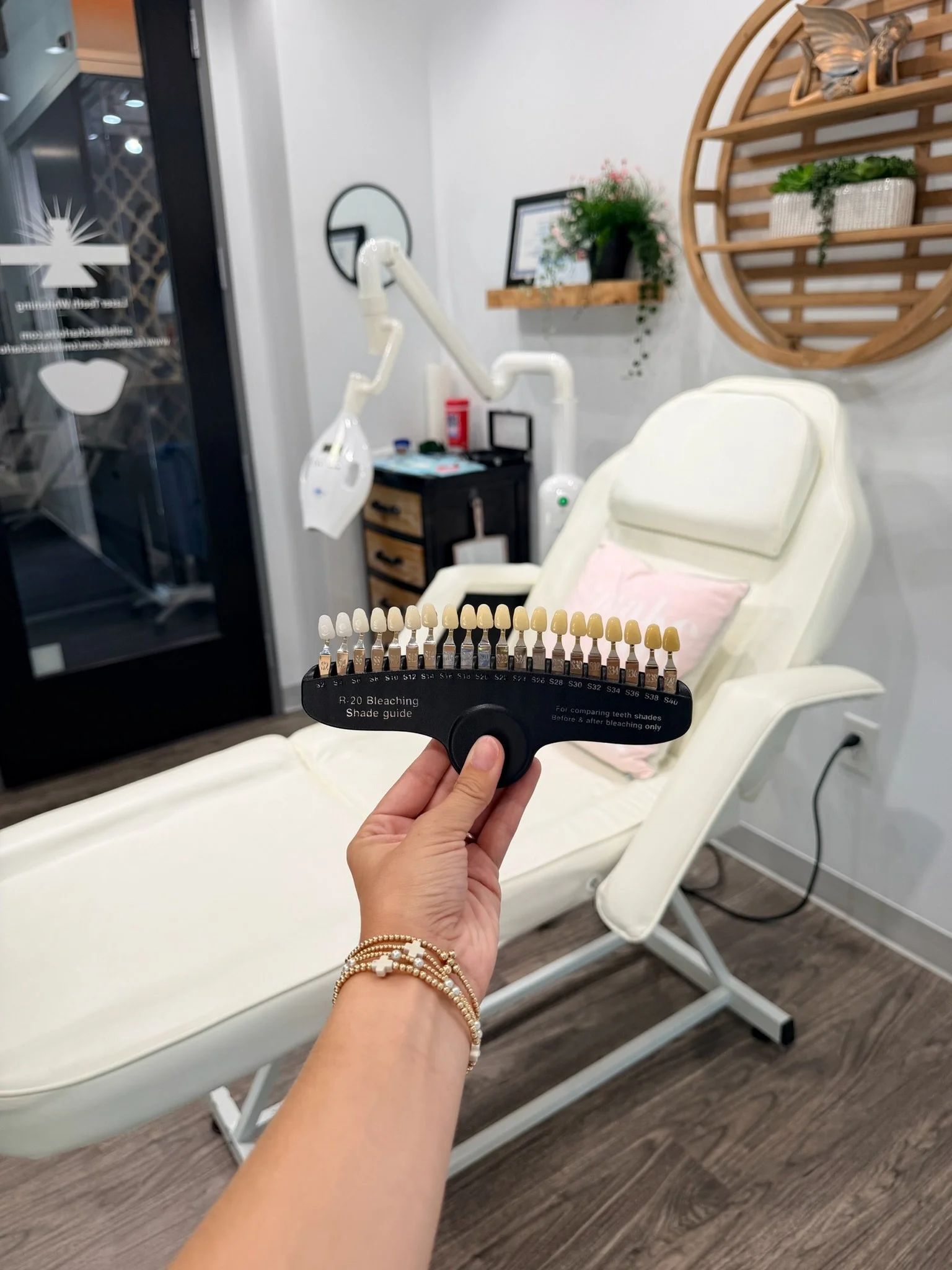 A woman's hand holding up a tooth shade range guide in a dental office with a white reclining chair in the background.