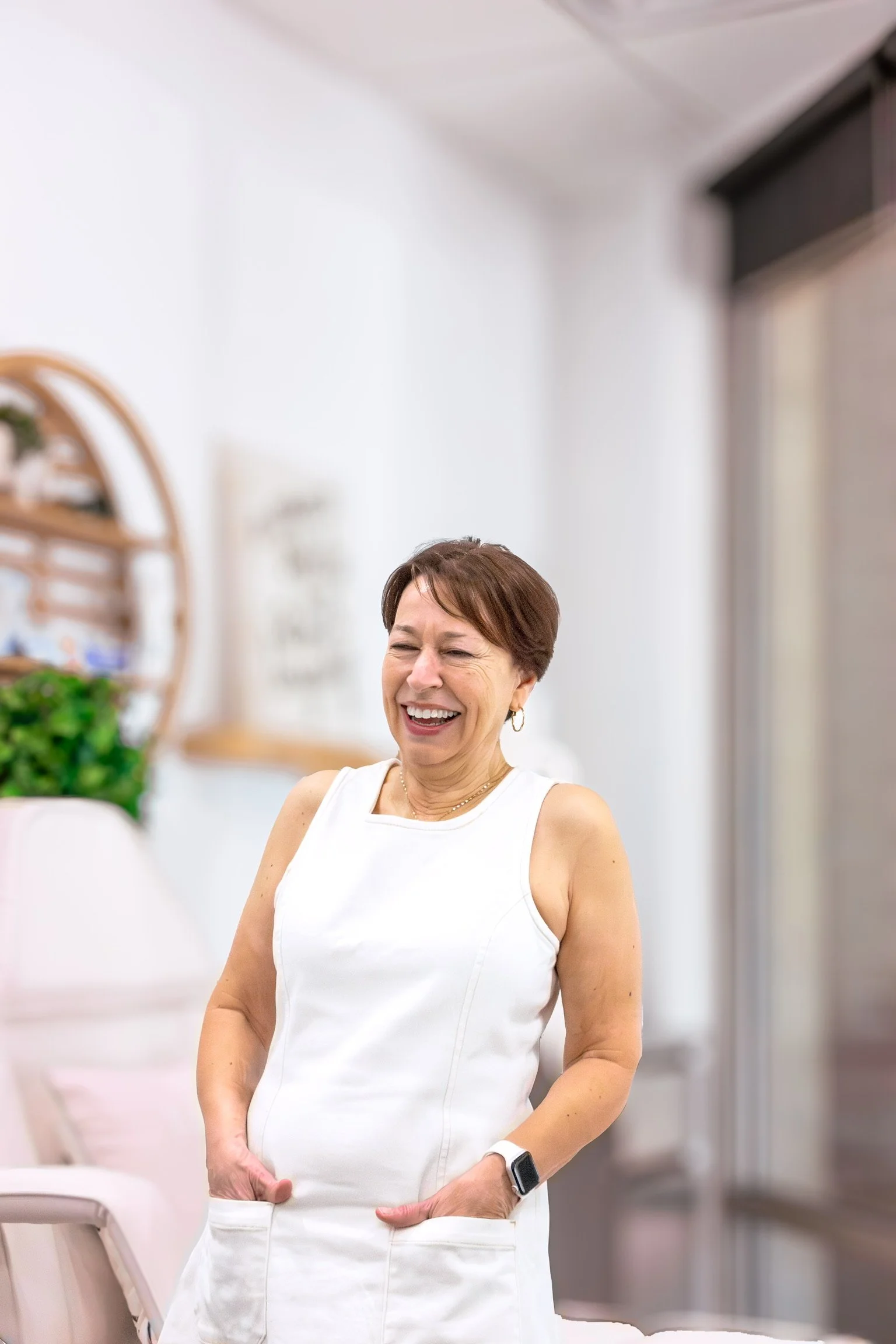 A woman (Chris) with short brown hair wearing a white sleeveless dress and a smartwatch on her left wrist, smiling and standing indoors.