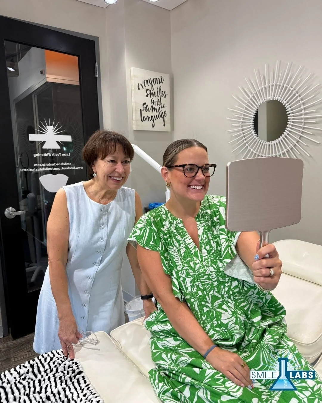 A woman in a green floral dress a mirror in a dental office, smiling. Chris is standing behind her, also smiling. The office has a decorative mirror on the wall, a sign that says 'Everyone smiles in the same language.'