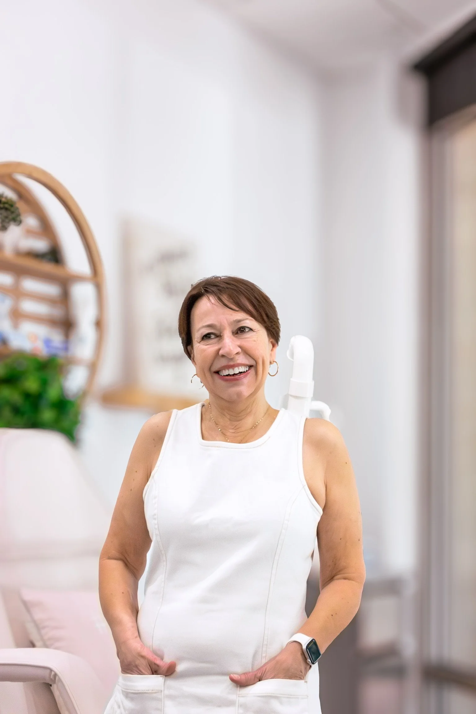 A smiling woman (Chris) with short brown hair, wearing a white dress, and standing in a well-lit room with white walls and furniture, and a wooden shelf with decorative plants in the background.
