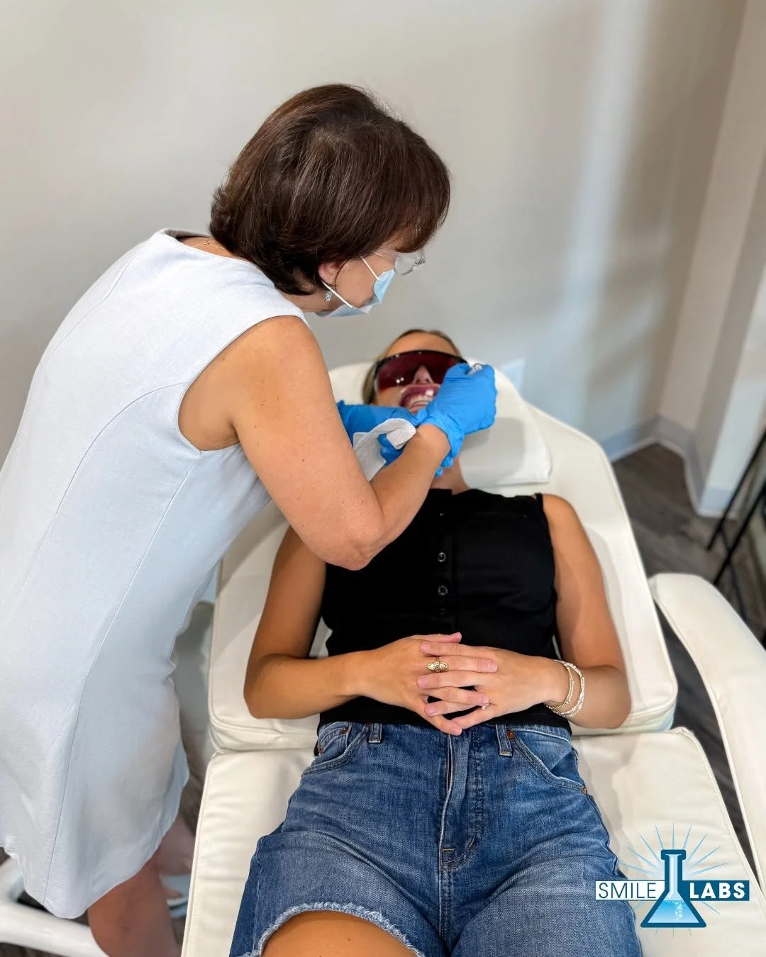 A woman lying on a medical chair receiving a dental procedure from a female healthcare professional wearing a mask, gloves, and protective glasses.