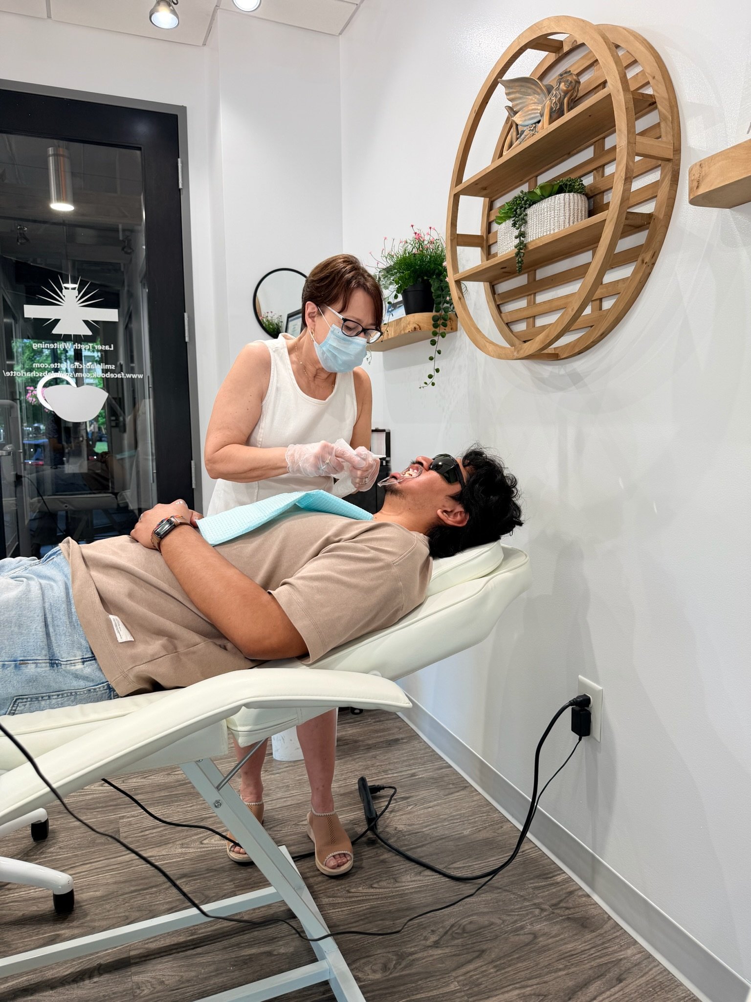 A man is lying on a dental chair receiving a tooth whitening treatment from a dental professional, wearing a mask and gloves.
