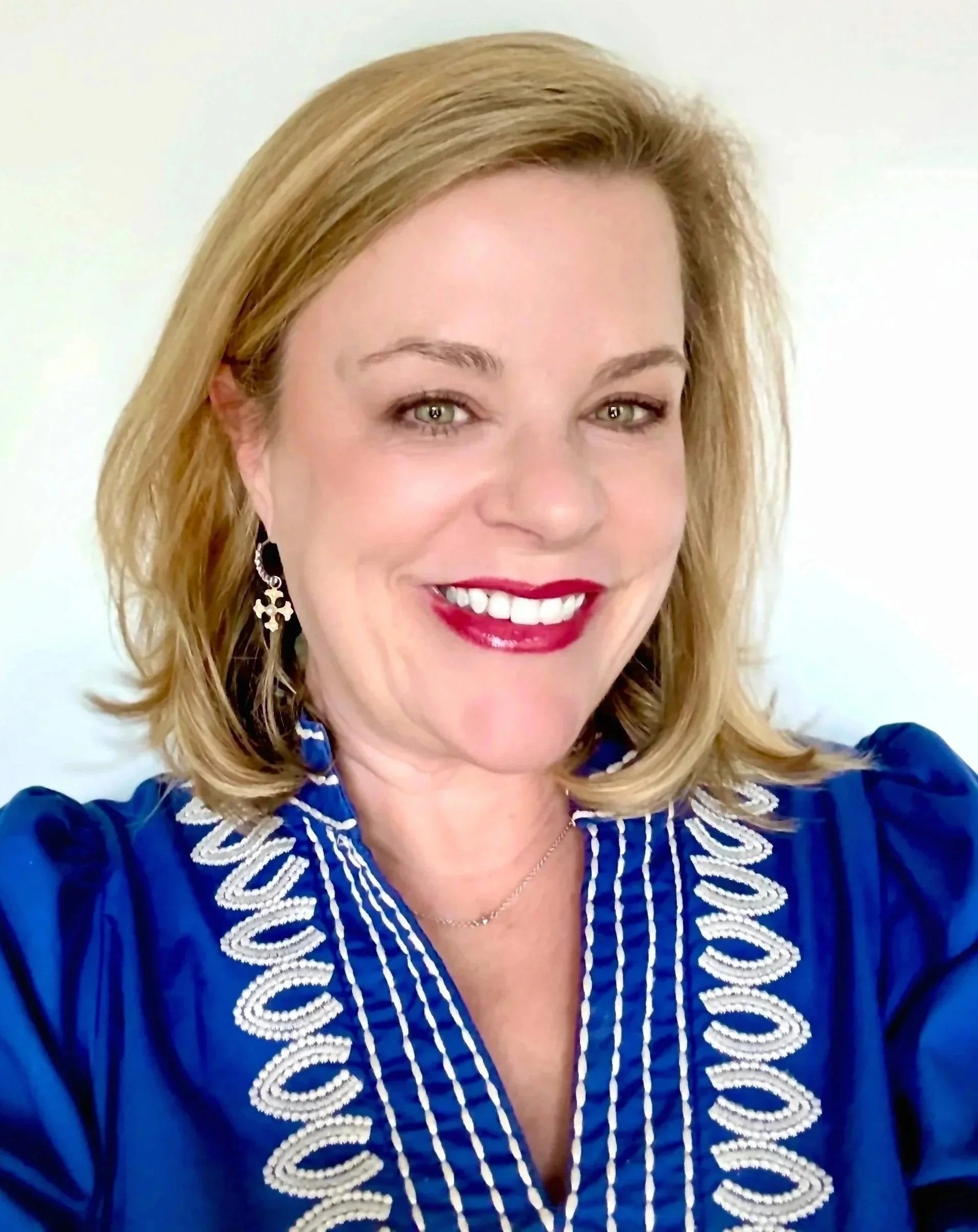 A woman with blonde hair, green eyes, and red lipstick smiling, wearing a blue dress with white embroidered details and a silver necklace, against a plain white background.
