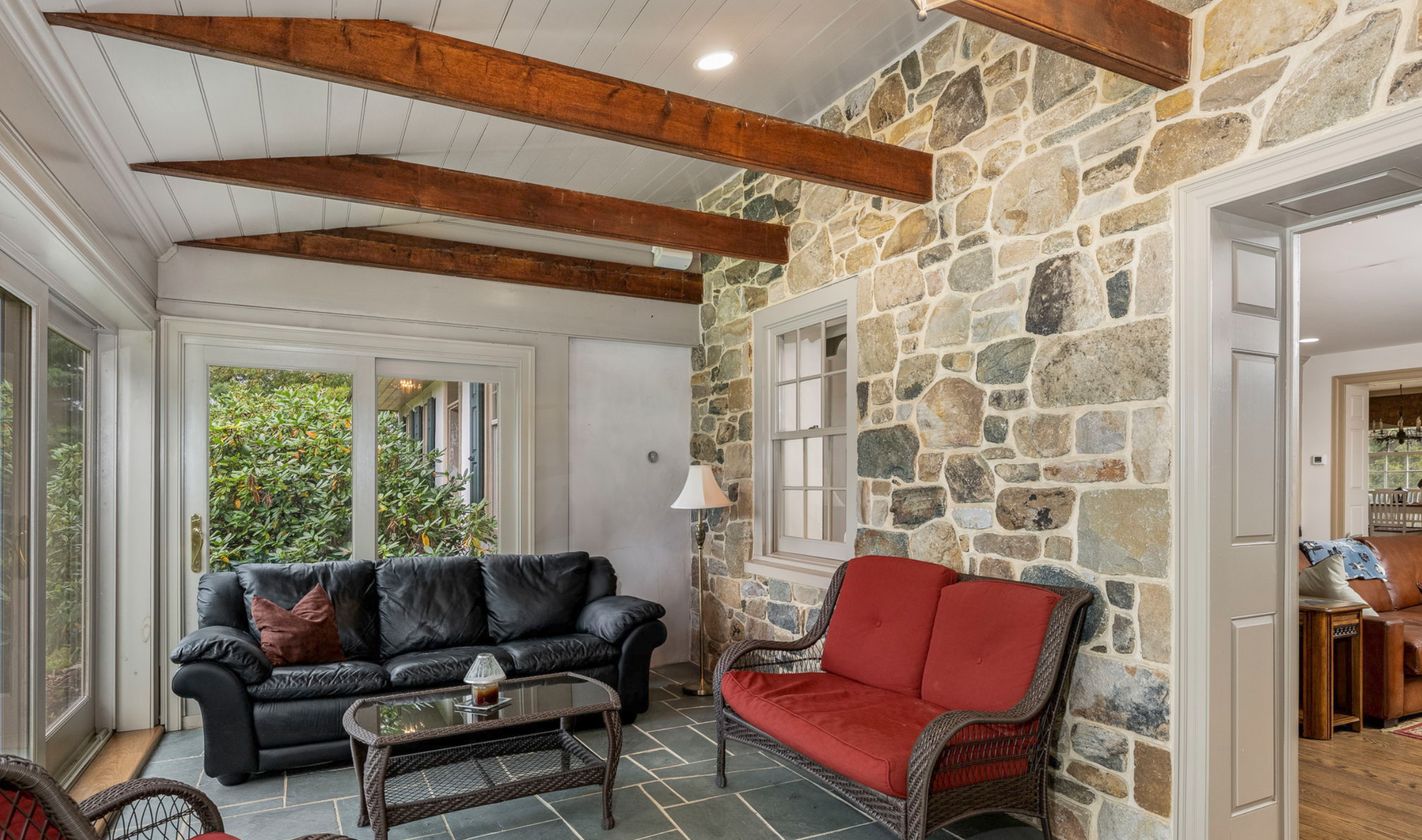 Living room with stone accent wall, wooden beams on the ceiling, leather and wicker chairs, and sliding glass door leading outside.