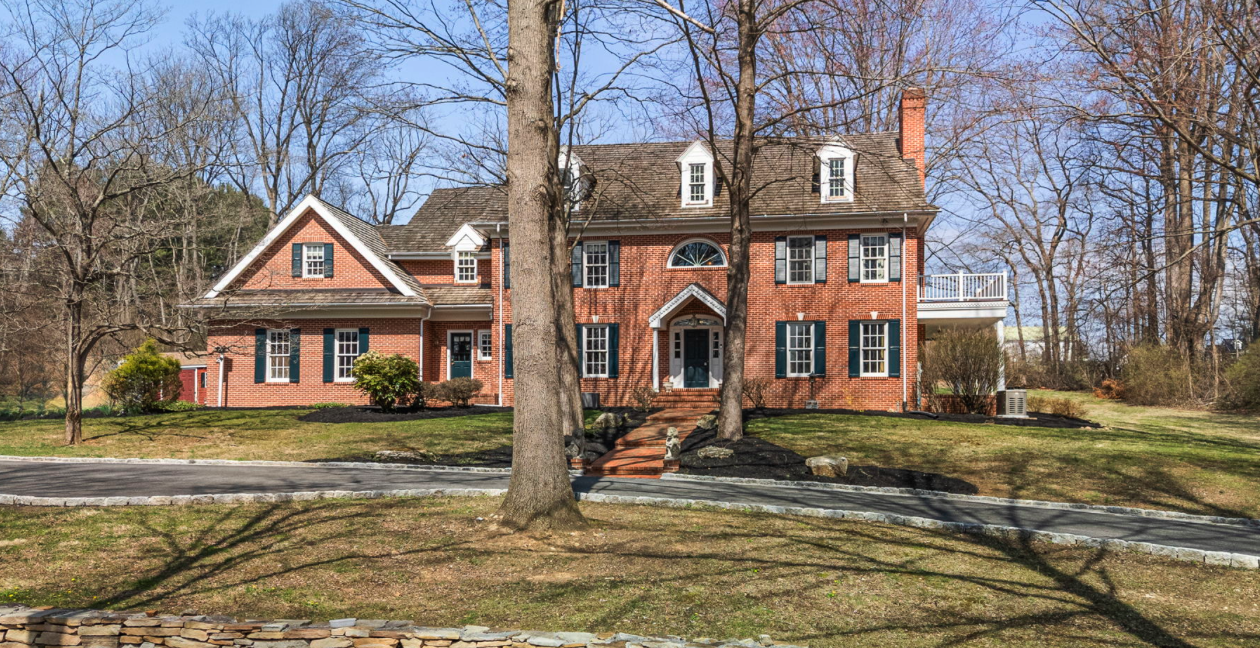 A large brick house with multiple stories, dormer windows, a front porch, and a driveway, surrounded by leafless trees and a lawn.