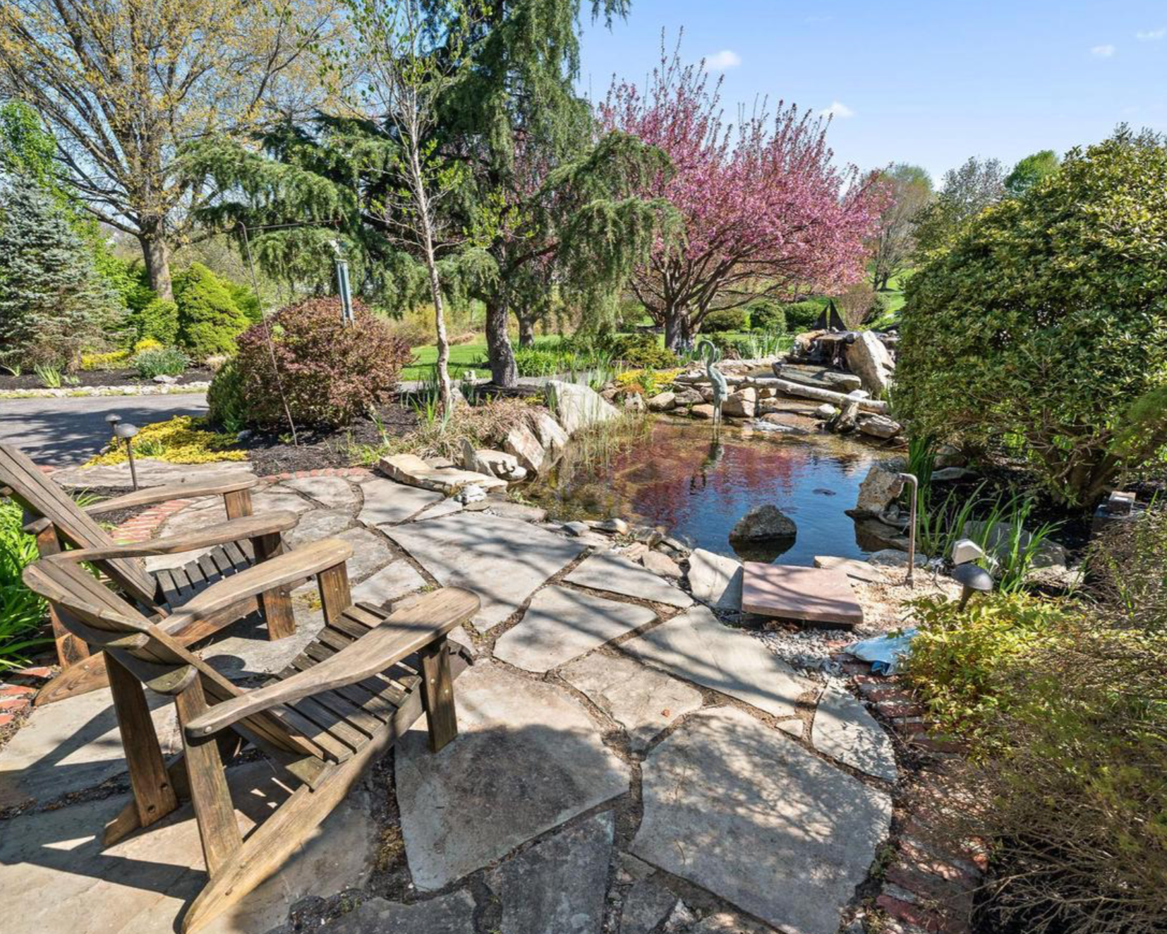 A backyard garden with stone pathway, wooden chairs, small pond, trees with pink and green foliage, and a clear blue sky.